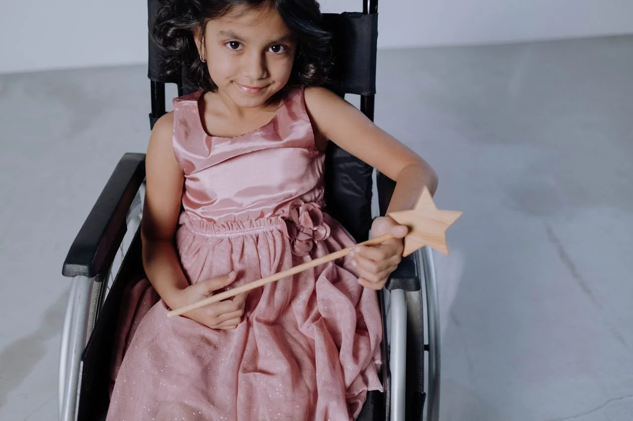 A young girl sitting in a wheelchair, wearing a pink satin dress, holding a star-shaped wand, and smiling at the camera.
