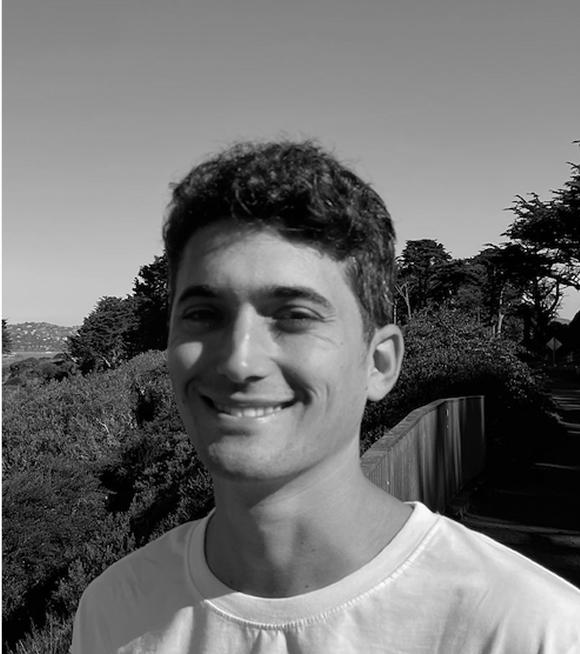 A young man with curly hair smiling outdoors with trees and a fence in the background, in black and white.