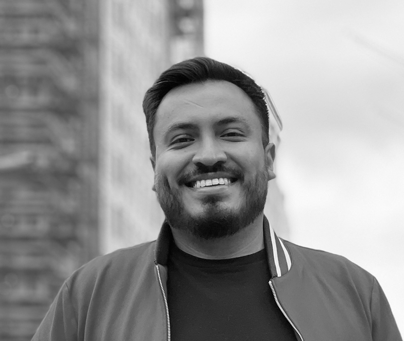 Smiling man with beard and short hair, wearing a jacket and t-shirt, standing outdoors near a brick wall.