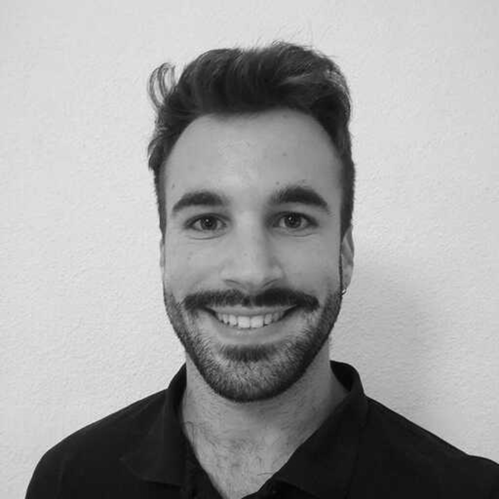 Black and white photo of a smiling man with dark hair and a beard, wearing a collared shirt, standing against a plain wall.
