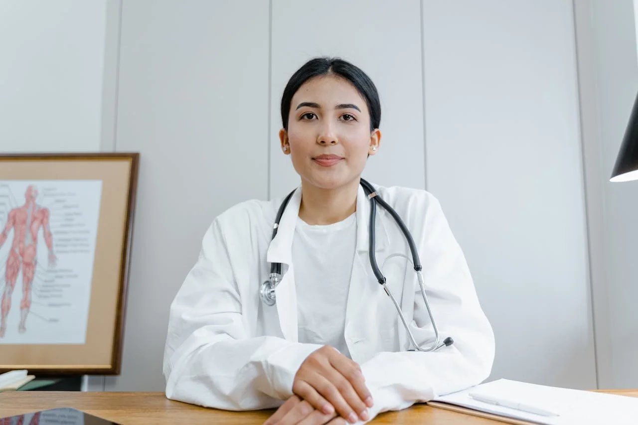 Female doctor sitting at a desk with a stethoscope around her neck, in a medical office with a human anatomy chart in the background.