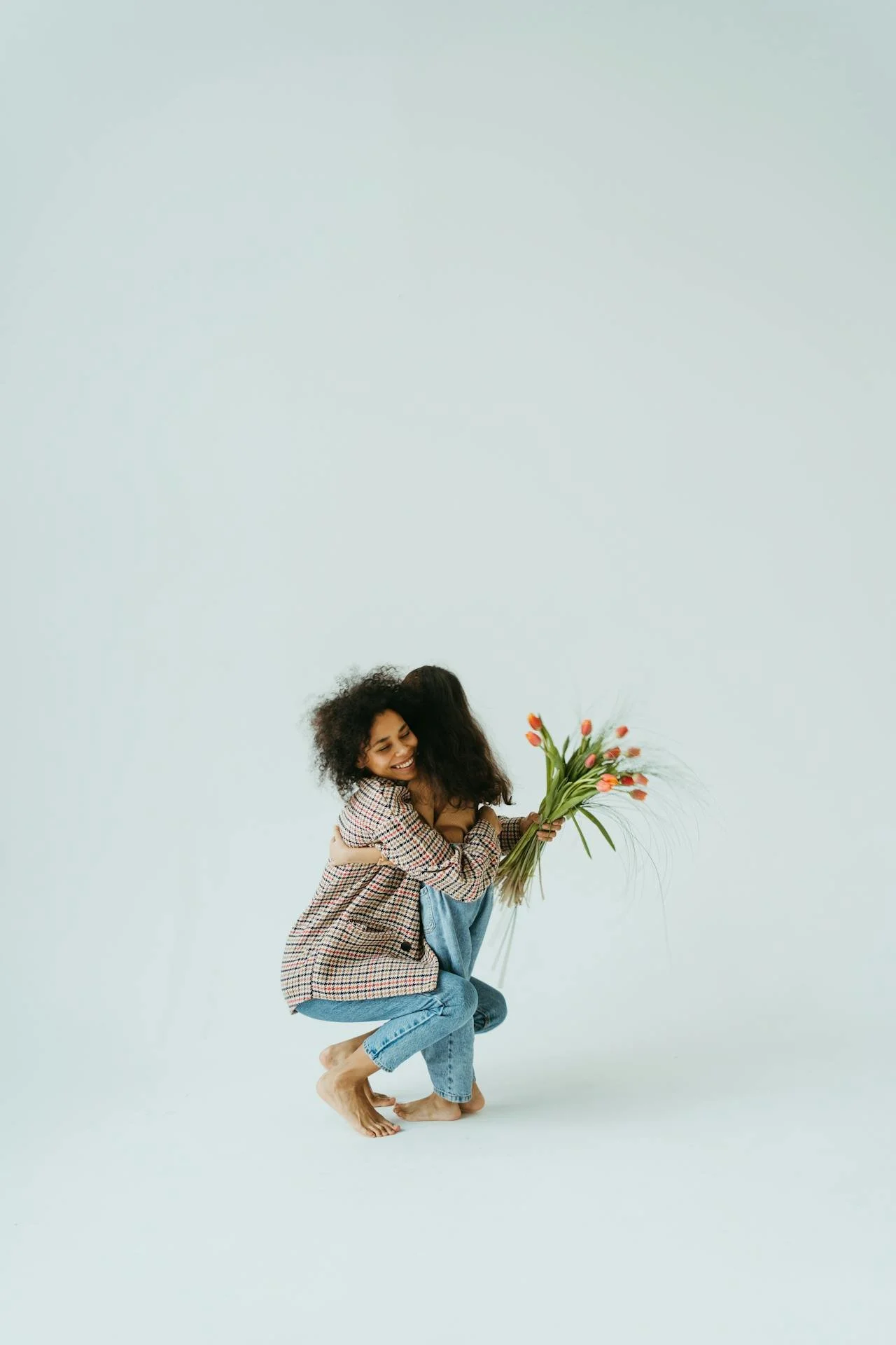 Two women hugging, one holding a bouquet of pink tulips, in a minimal studio setting.