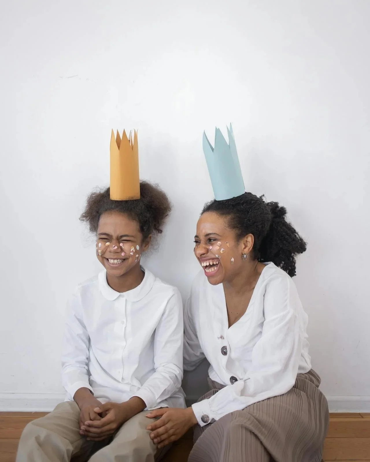 Two women sitting on the floor against a white wall, both wearing paper crowns and smiling or laughing, with face decorations.
