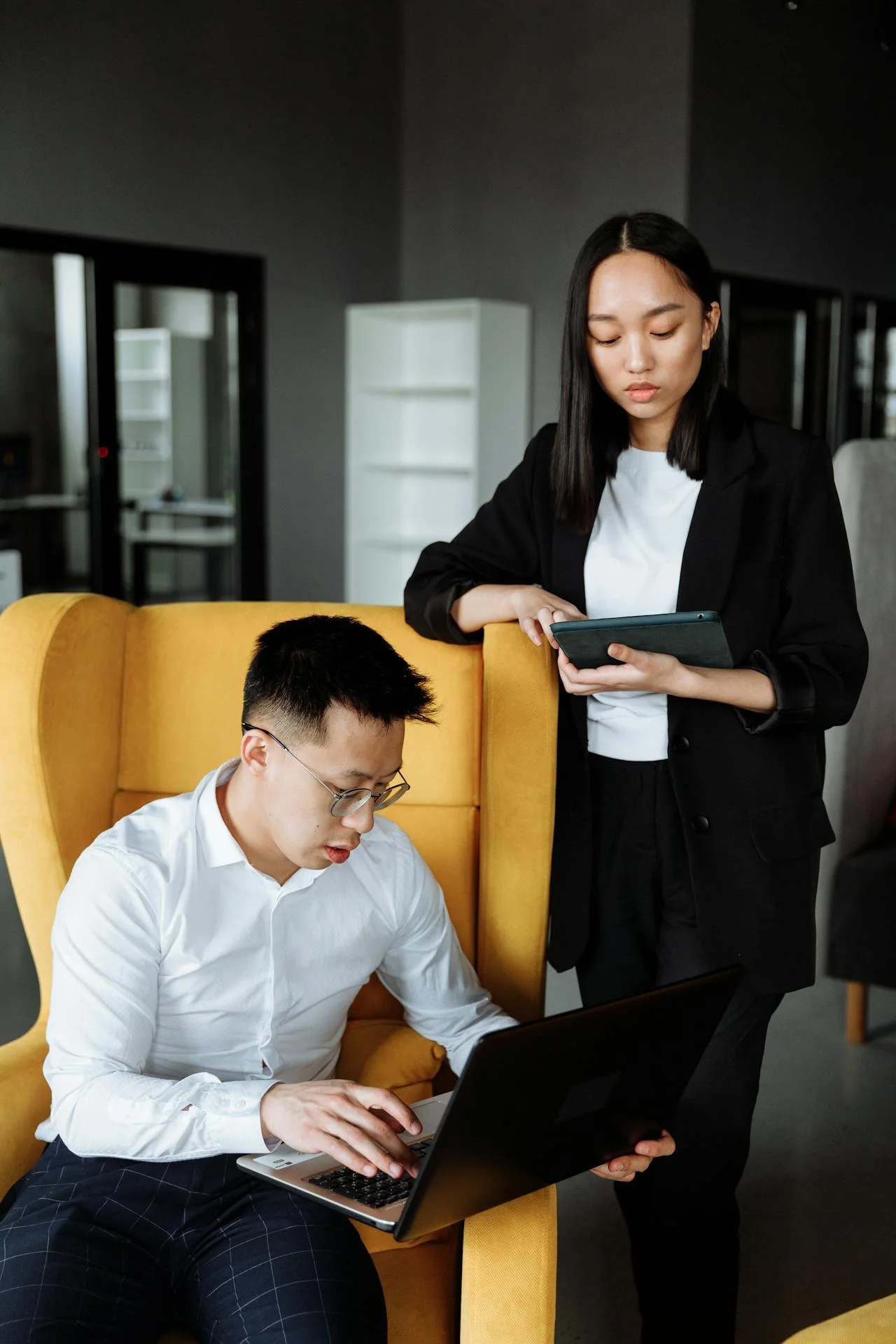 A man sitting in a yellow chair working on a laptop, with a woman standing next to him holding a tablet, in a modern office setting.