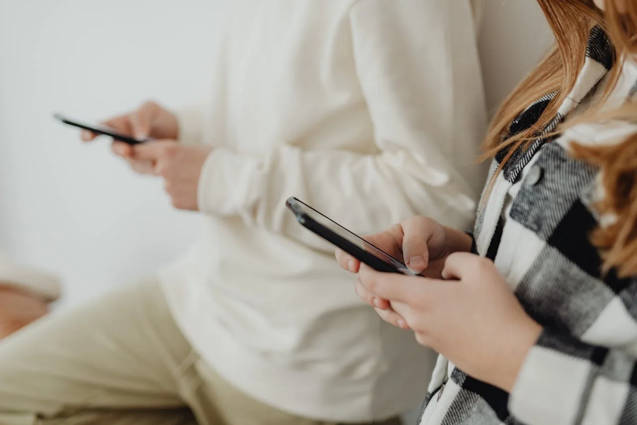 Two people sitting and using their smartphones. One person wears a white shirt, and the other wears a plaid shirt.