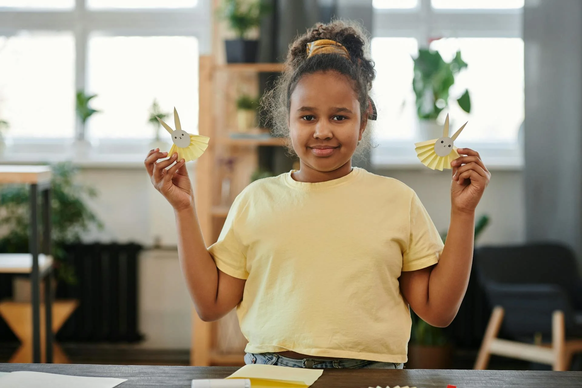 A young girl holding two paper bunny crafts in an indoor setting with plants and large windows in the background.