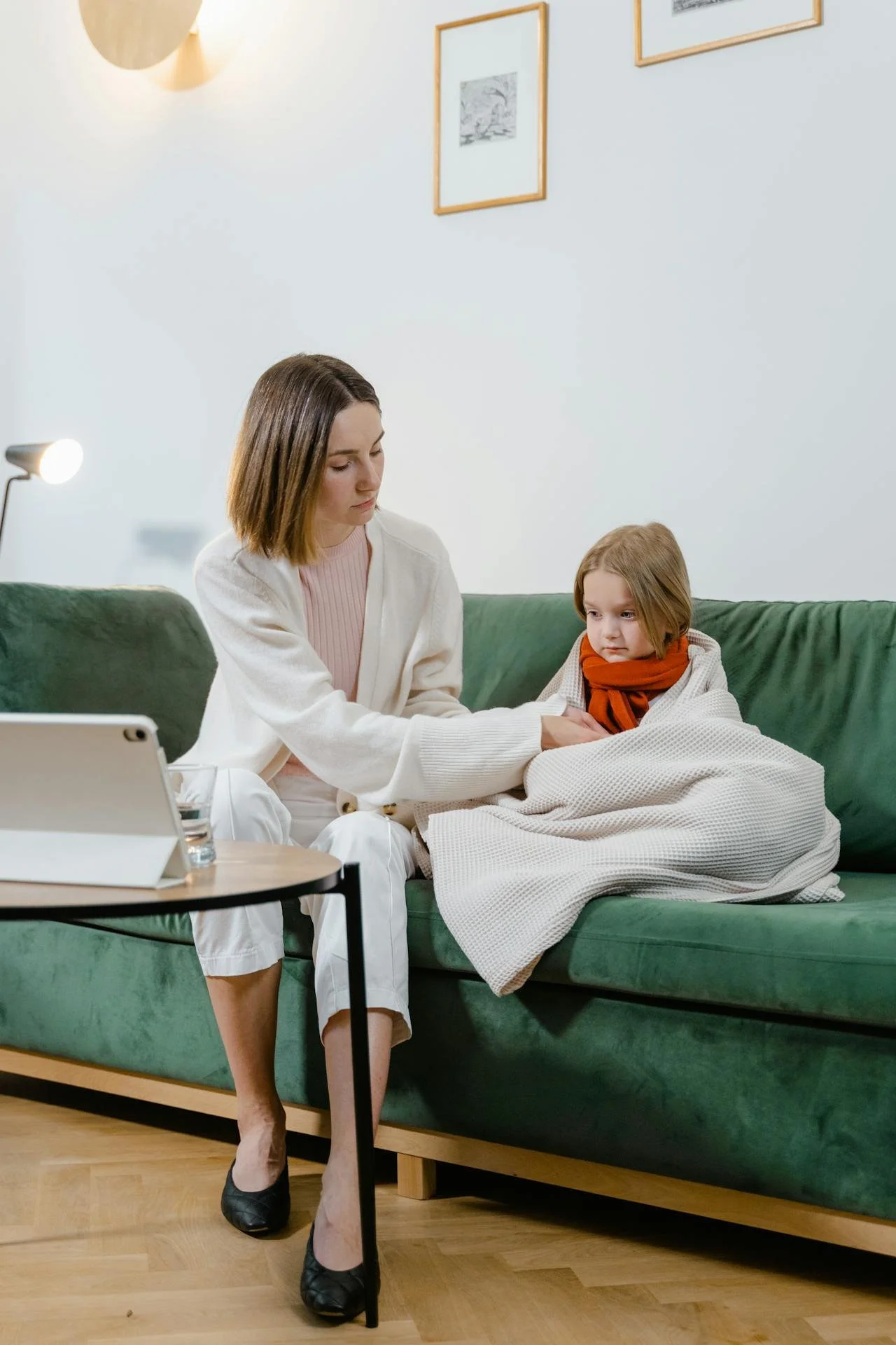A woman sits on a green sofa with a young girl. The woman appears to be comforting the girl, who looks sad or upset. There is a tablet and a glass of water on a small side table in front of them.