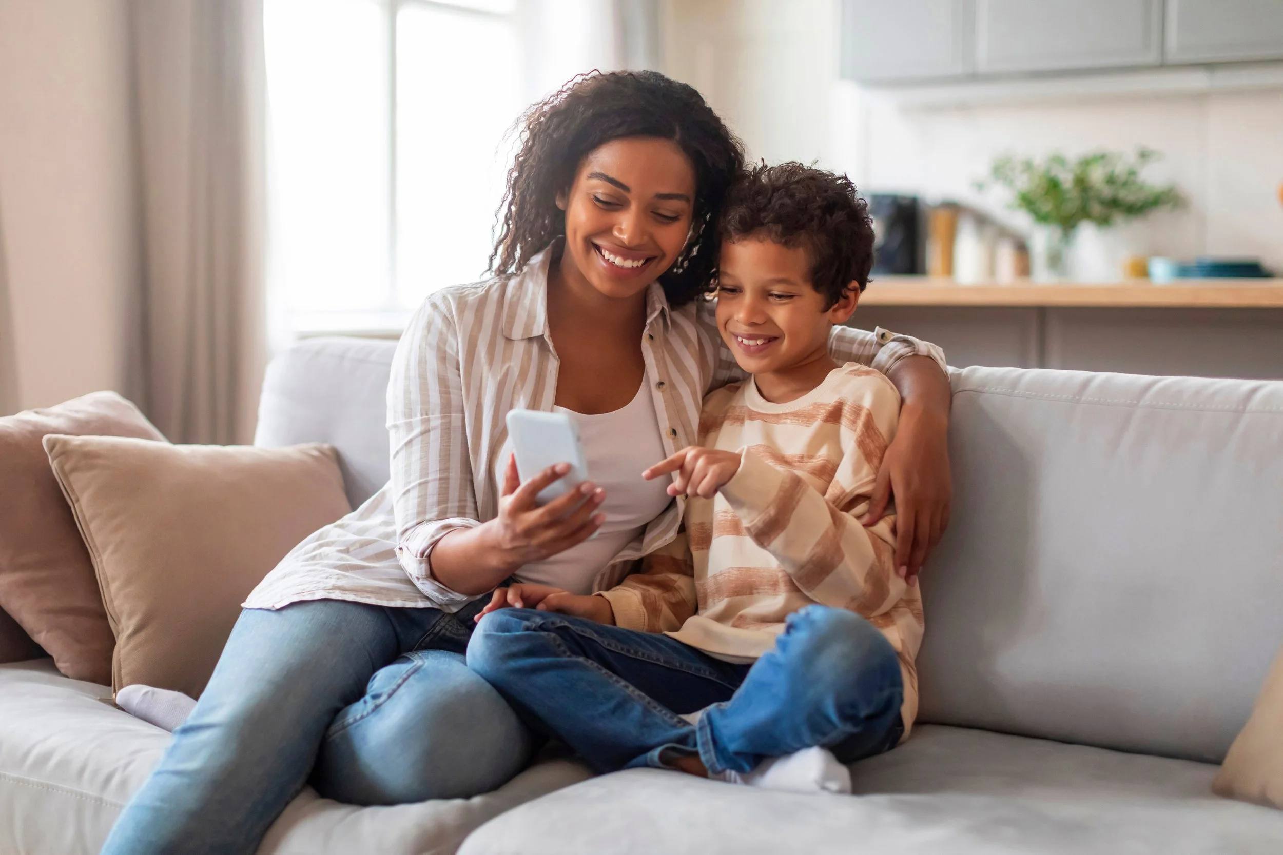 A smiling woman and boy sit on a white couch, looking at a phone together in a bright living room.