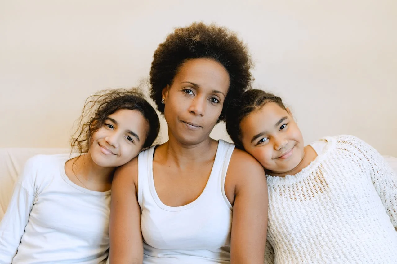 A woman sitting on a couch with two young girls, all smiling softly, with light-colored background, casual white tops, one wearing a knit sweater.