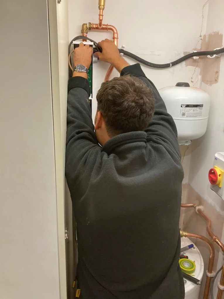 A man working on plumbing pipes in a utility room, with copper and black pipes visible on the wall.