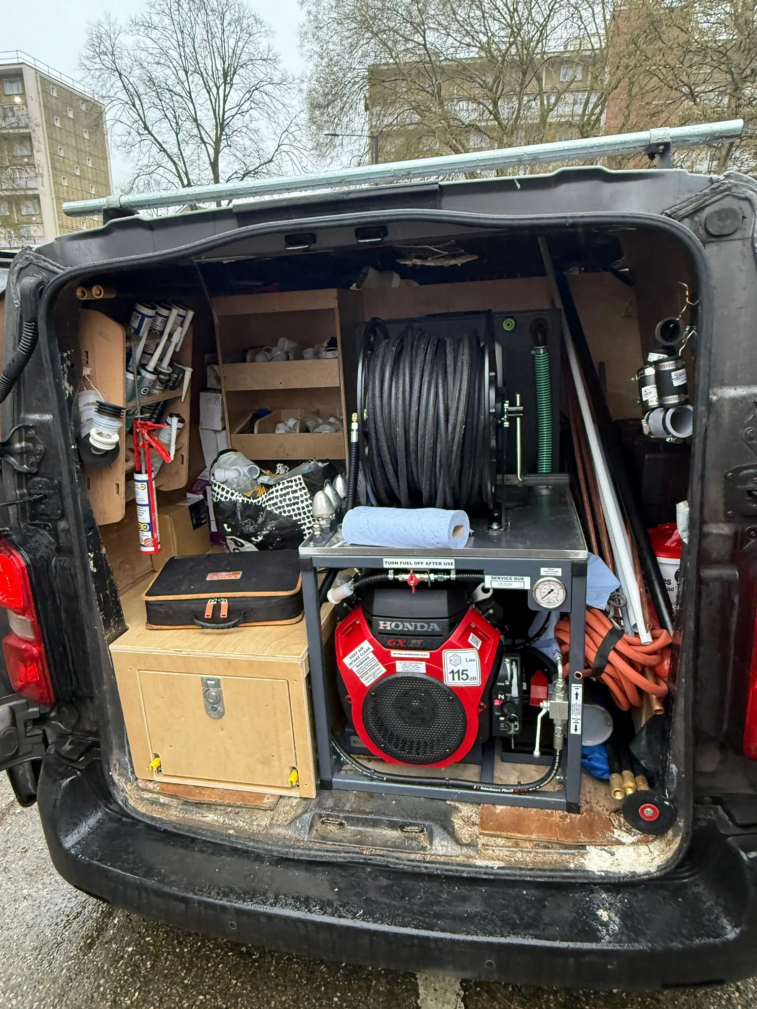 Rear of a van with equipment, including a Honda gas generator, hoses, tools, and storage shelves for plumbing or maintenance work.