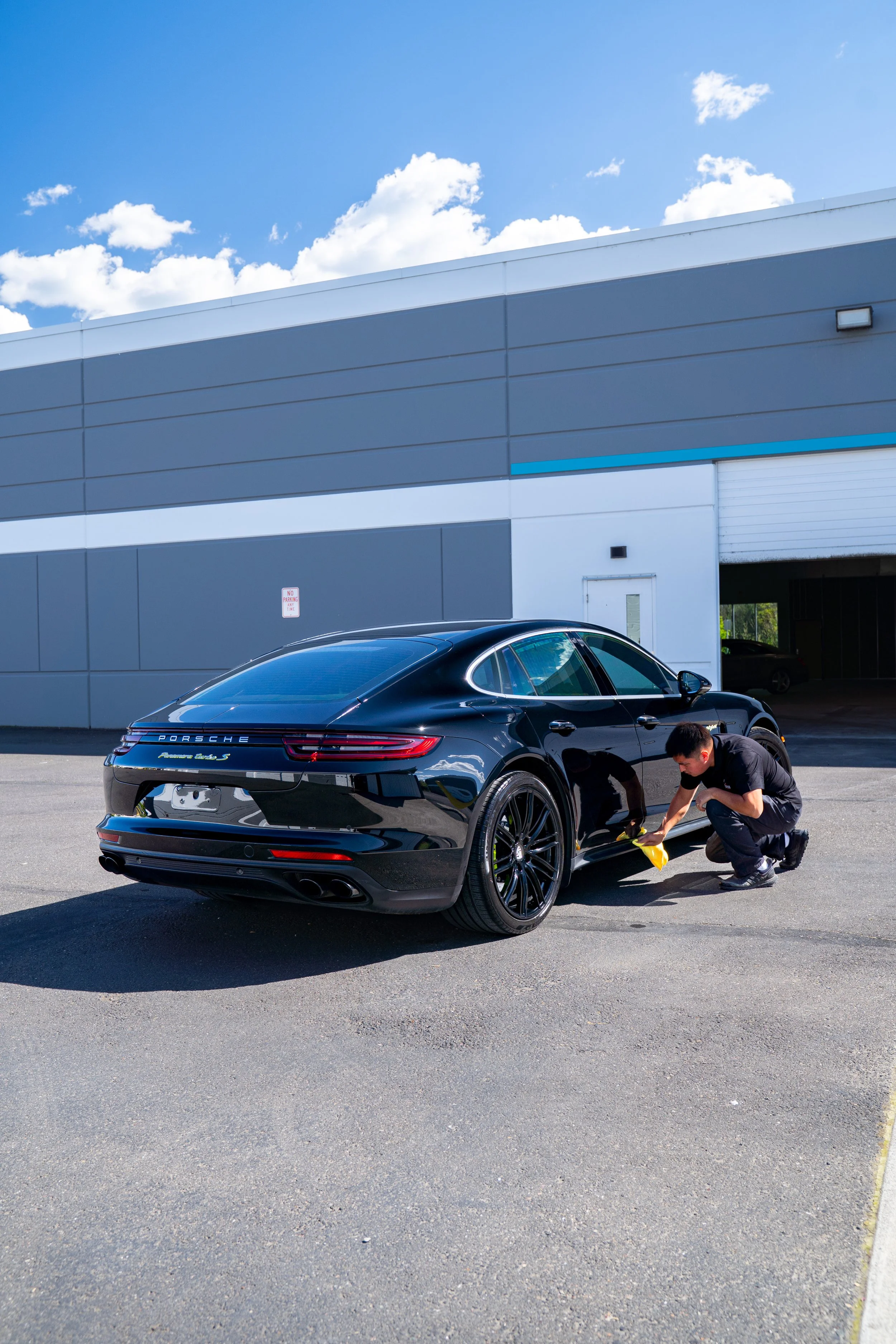 A man crouching beside a black Porsche car in a parking lot, working on the front left wheel, with a large gray commercial building and blue sky with clouds in the background.