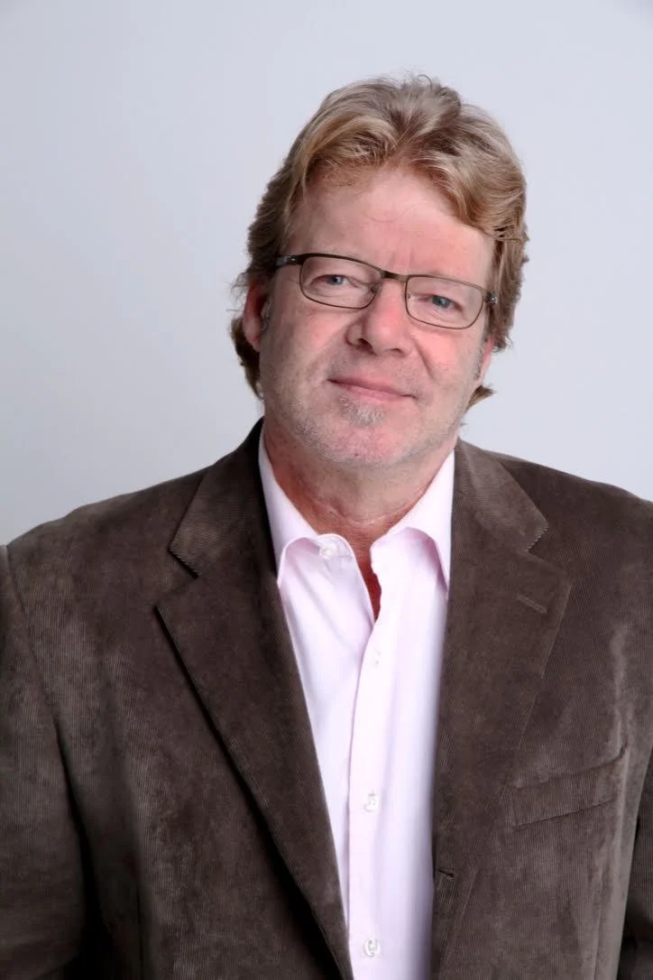 Blond haired man wearing glasses and a brown suit jacket.