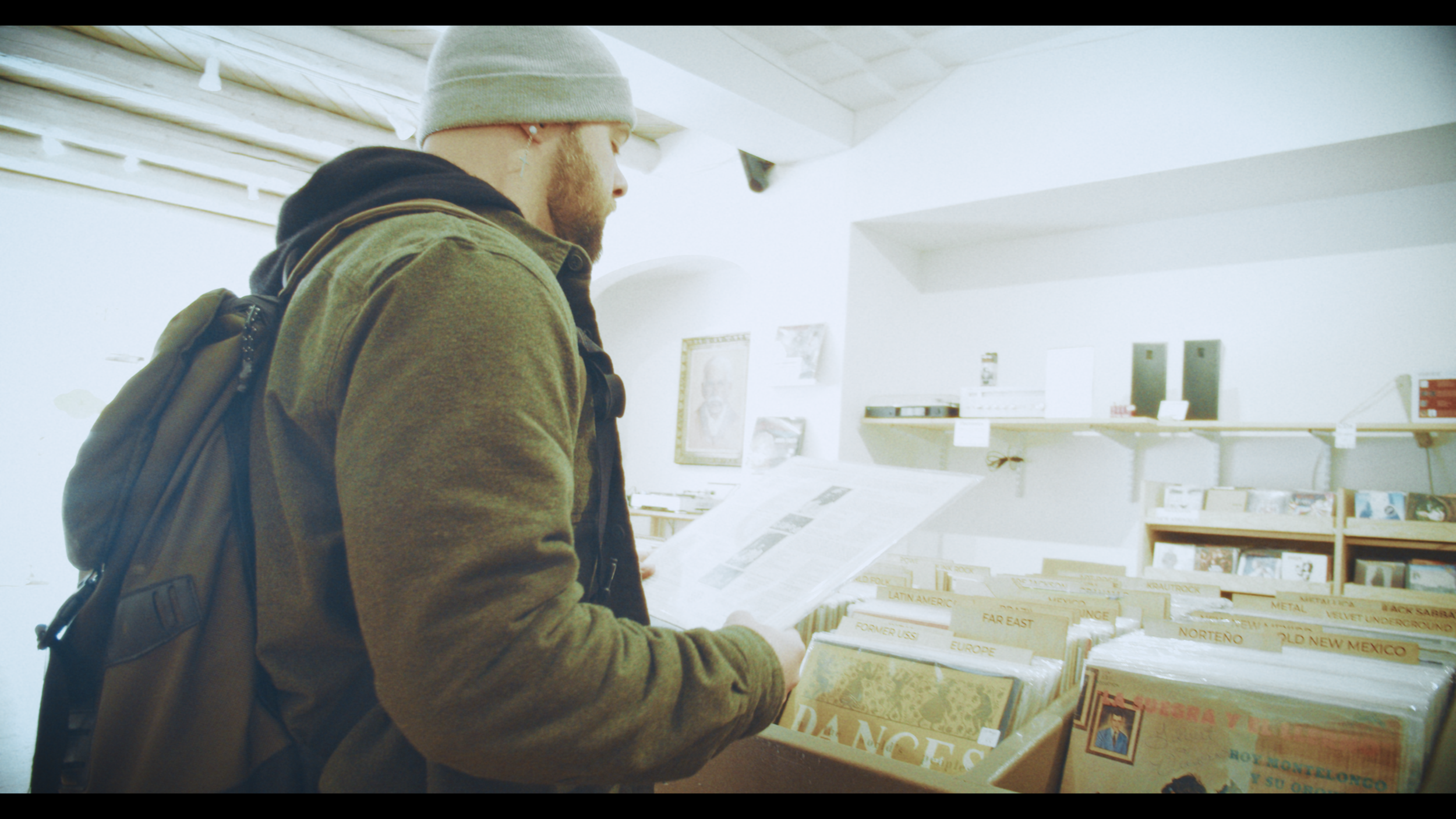 A young man in a gray beanie and brown jacket with a backpack is standing in a record store, holding and reading a newspaper or flyer. The store has shelves with records labeled by regions such as Europe, Far East, and Mexico.