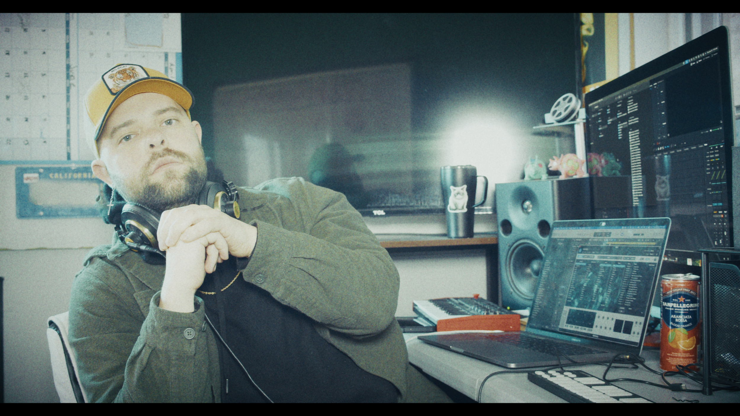 A man with a beard wearing a yellow cap, headphones around his neck, sits in a music production studio with multiple computer monitors, audio equipment, and a laptop, looking at the camera with a serious expression.