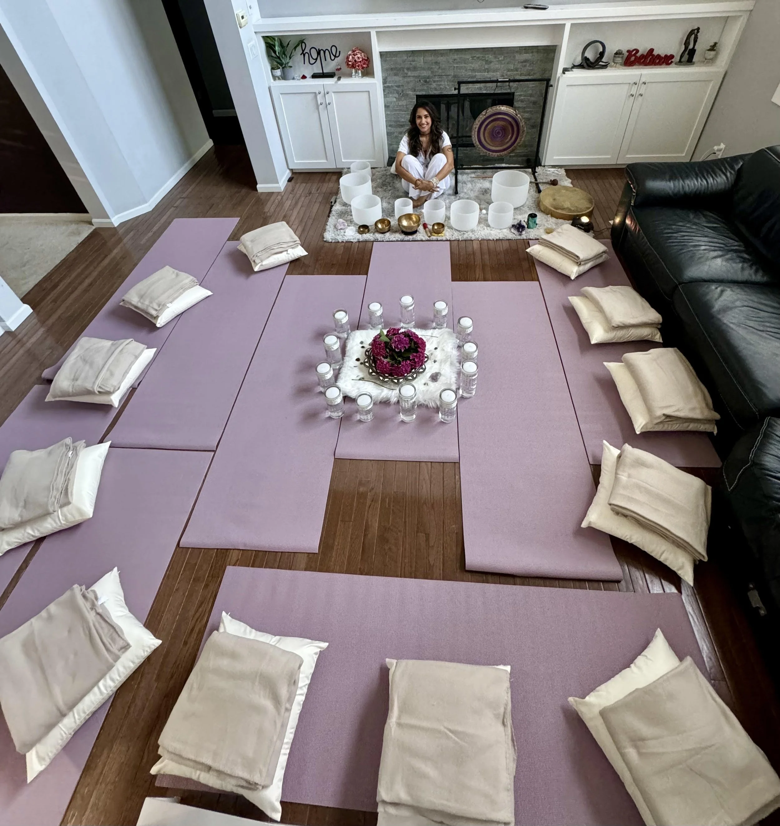 A woman sitting cross-legged on a blue mat with candles and flowers arranged for a meditation or spiritual session in a living room with pink mats and beige pillows.