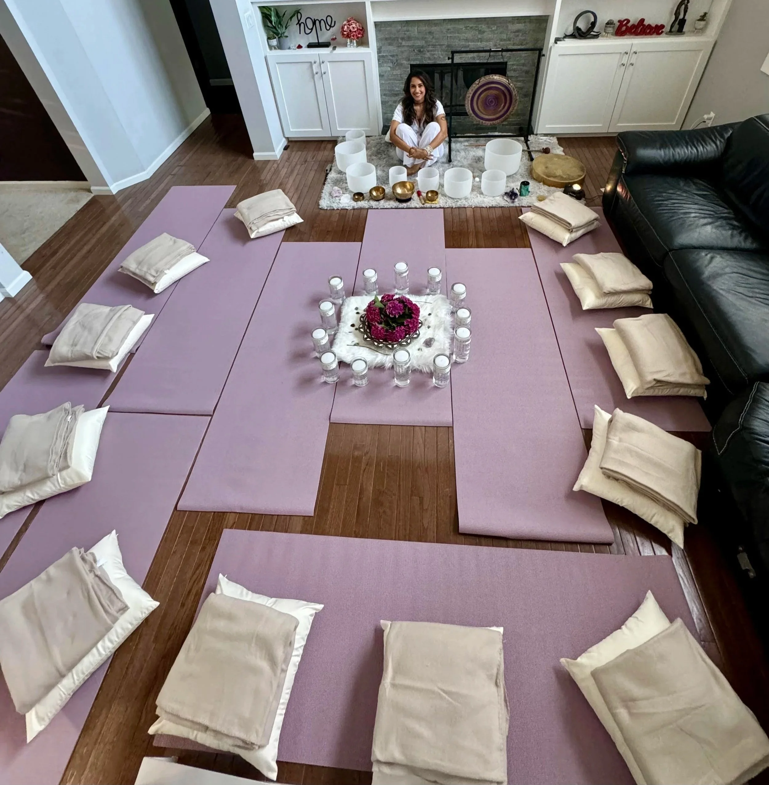 Living room with pink yoga mats arranged in a square for a group meditation or yoga session, with folded beige blankets and pillows on each mat. A woman in white sits cross-legged on the floor behind the mats, surrounded by white candles and decorative items, with a fireplace and shelving unit in the background.