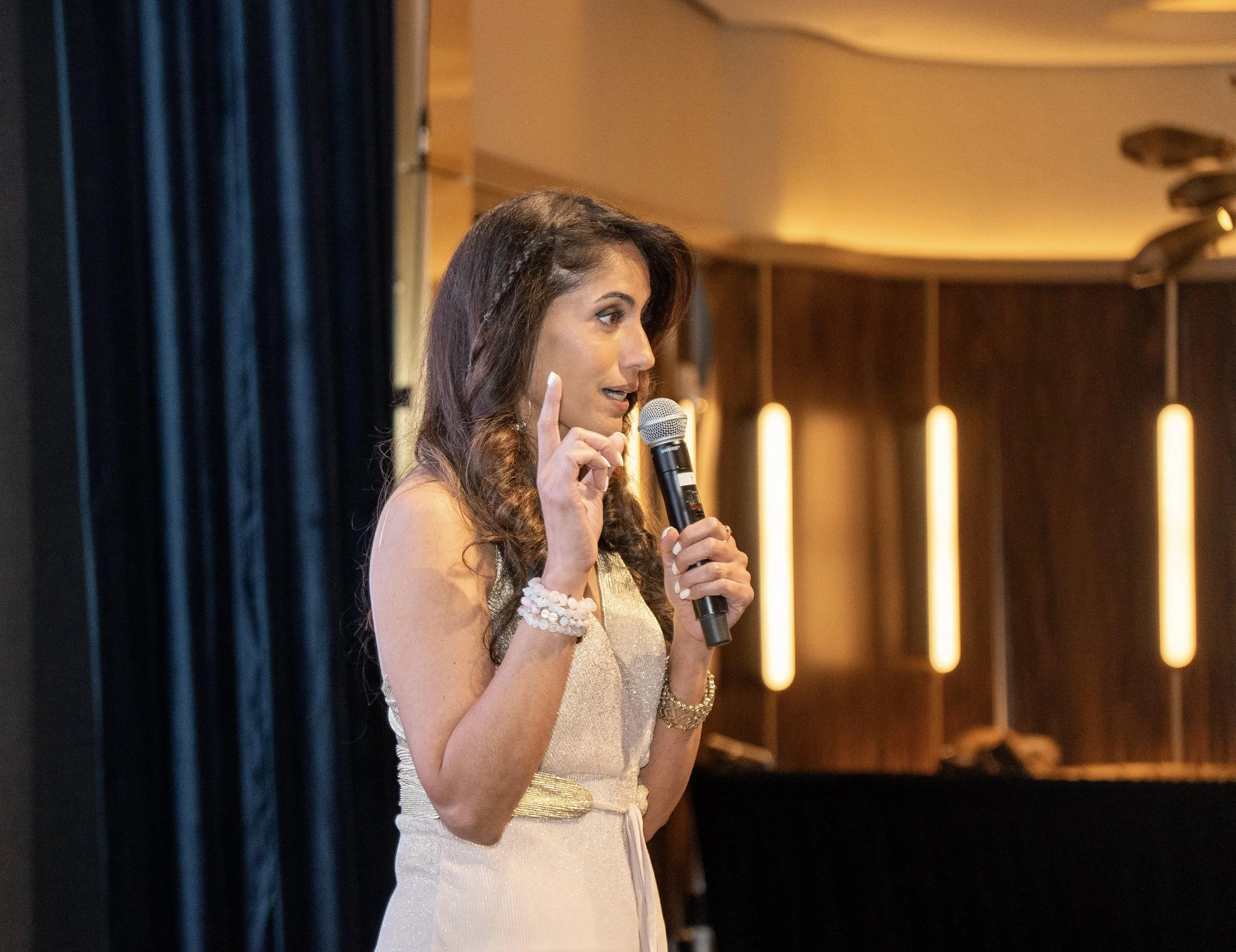 A woman with long brown hair holding a microphone, gesturing with her finger, speaking on stage in an indoor event space.