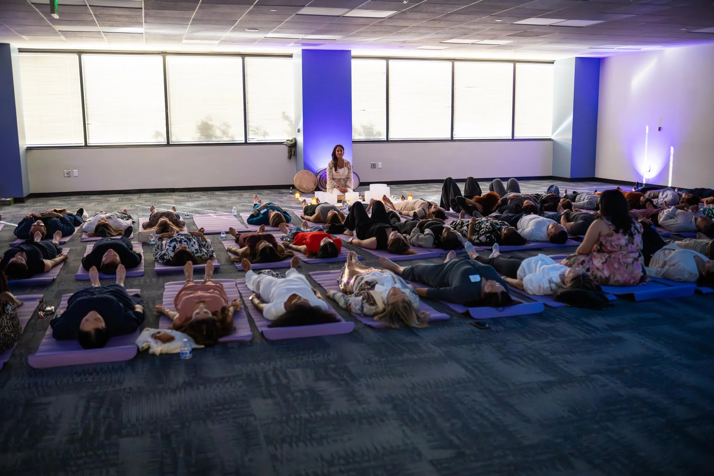 A large group of people participating in a yoga or meditation session in a spacious, well-lit room with large windows. Many individuals are lying on yoga mats, and a woman is sitting cross-legged at the front near candles and a drum.
