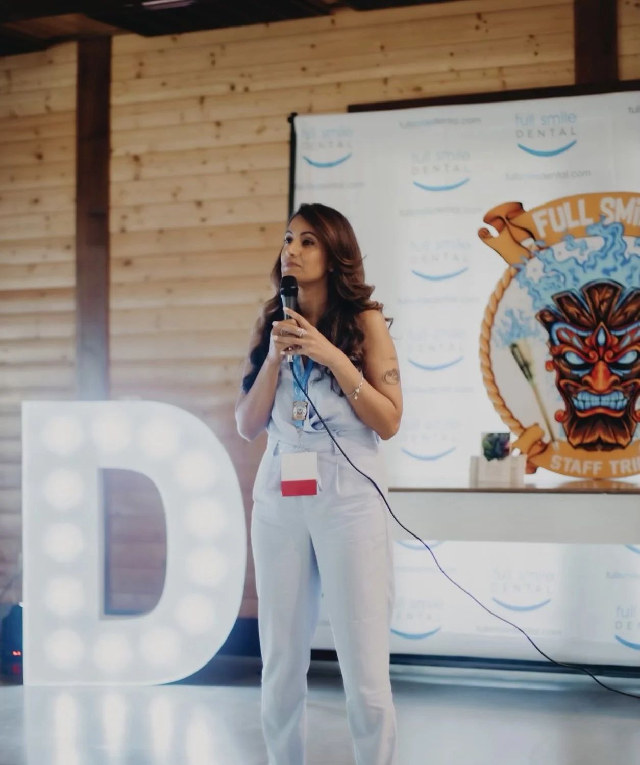 A woman with long brown hair holding a microphone stands in front of a banner with a colorful fierce mask logo. She wears a white outfit and a lanyard with a badge. To her left, a large illuminated letter D is visible.
