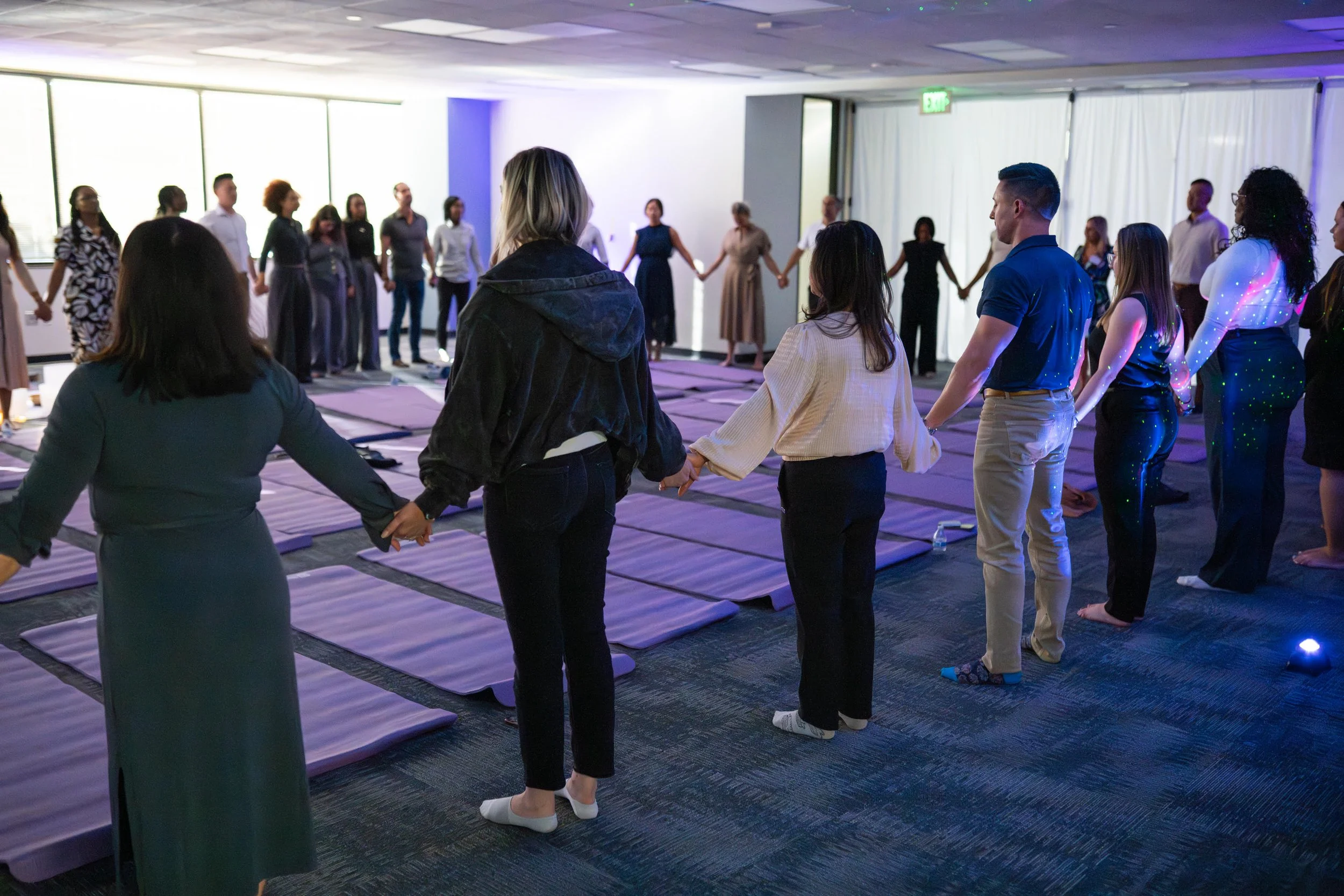 People holding hands in a circle during a group prayer or meditation session in a well-lit room with large windows and purple lighting.