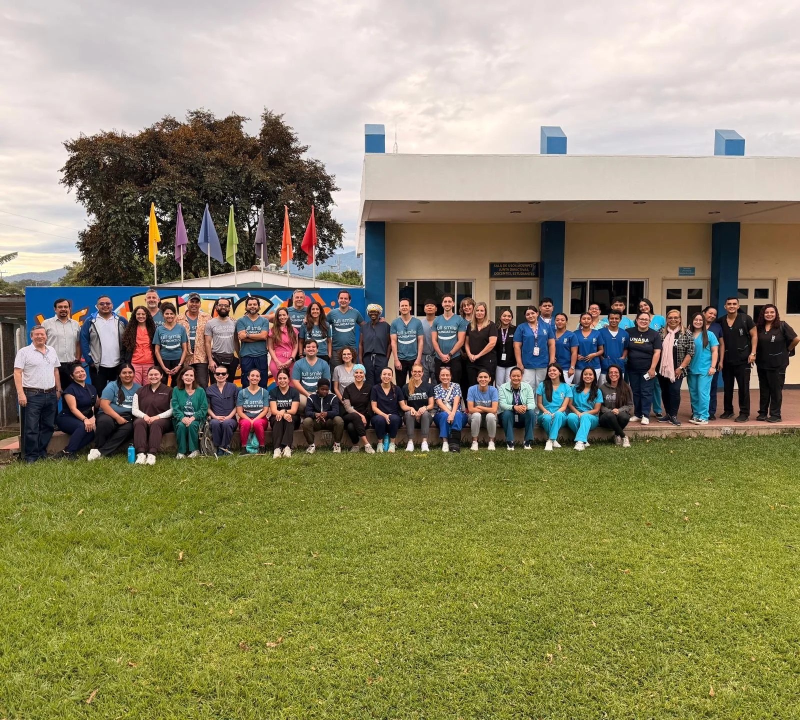 Group of people gathered outside a building with flags on poles, some wearing medical scrubs and others in casual attire, posing for a group photo.