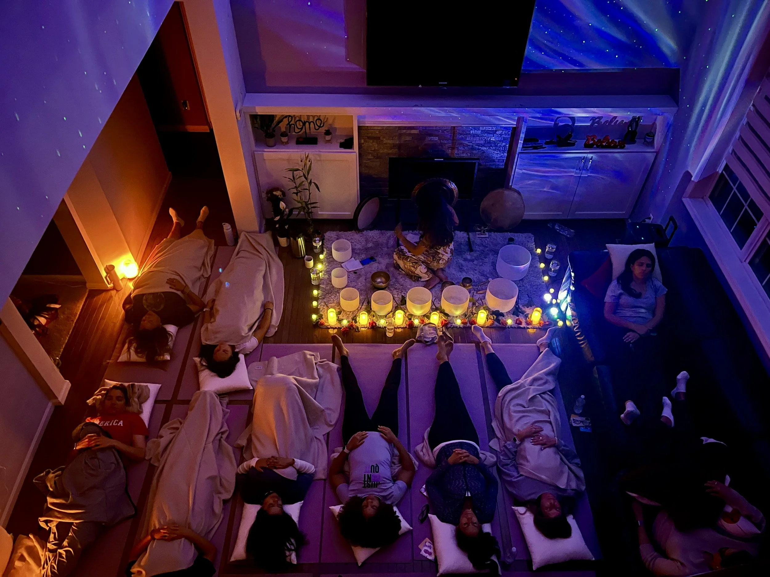 A group of people lying on mattresses on the floor in a living room during a meditation or relaxation session, with candles and colorful lights creating a calming atmosphere.