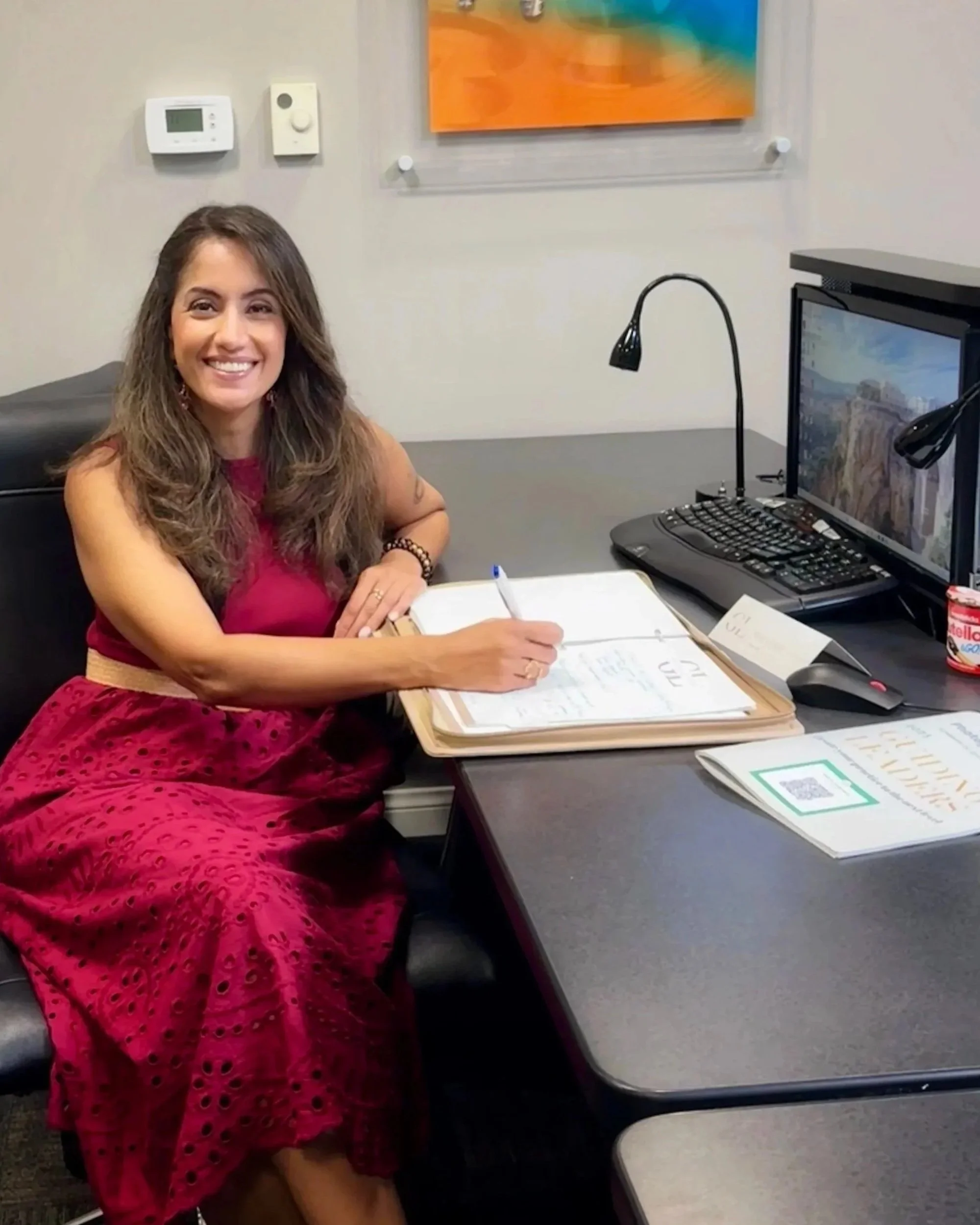 A woman sitting at a desk, smiling, writing in a notebook. She is wearing a red dress and is surrounded by office supplies, including a computer, mouse, desk lamp, papers, and a container of Nutella. There is a colorful painting and a thermostat on the wall behind her.