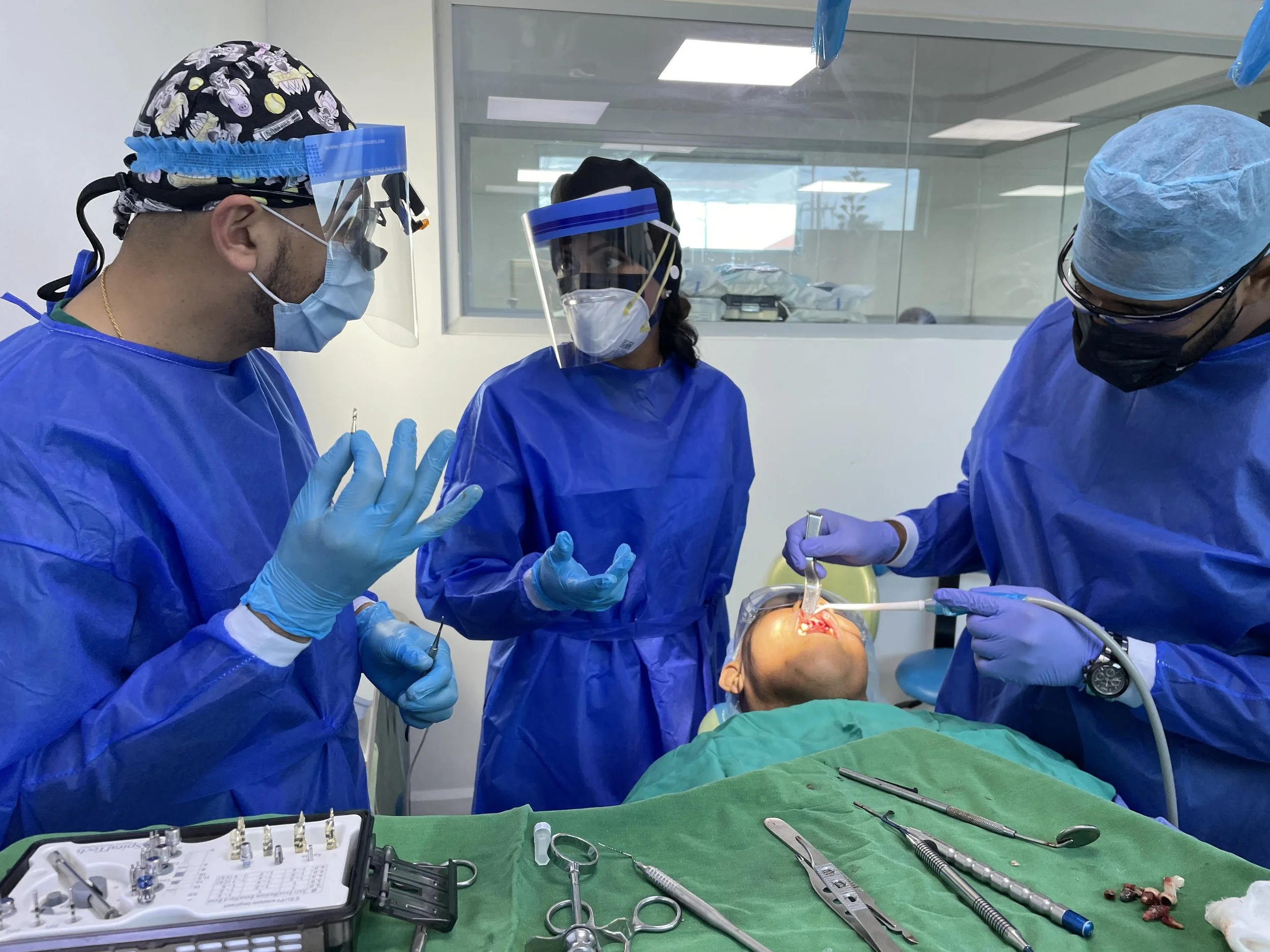 Three dental professionals in blue gowns, masks, and face shields performing a dental procedure on a patient lying on a dental chair in a clinical setting.