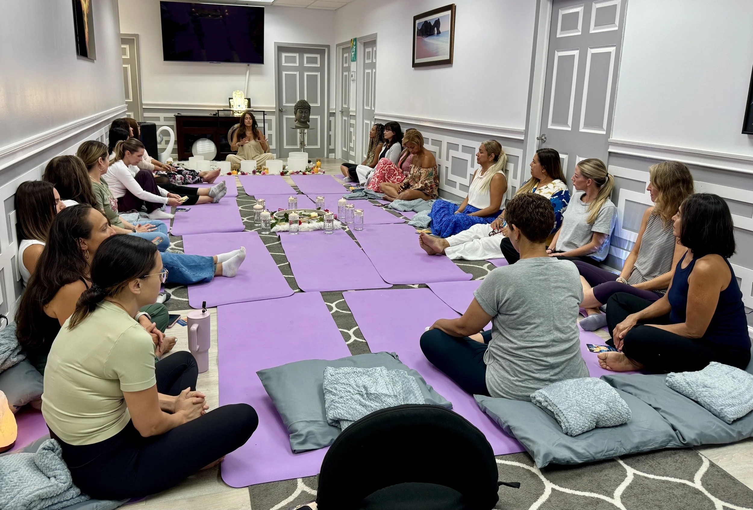 Group of people seated on purple yoga mats in a circle, participating in a meditation or breathing session in a well-lit, decorated room.