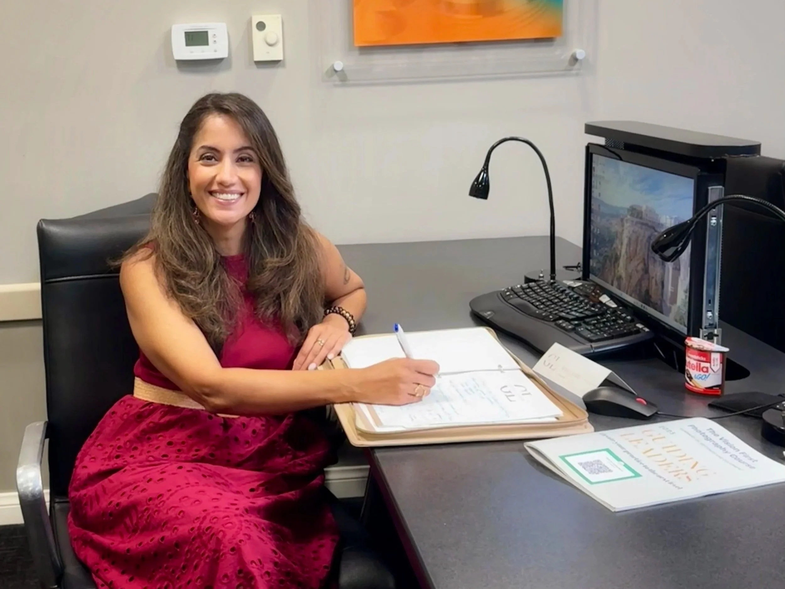 A woman with long brown hair wearing a red dress is sitting at a desk, smiling at the camera, with a pen in her hand and a notebook open in front of her, in an office setting with a computer and various items on the desk.