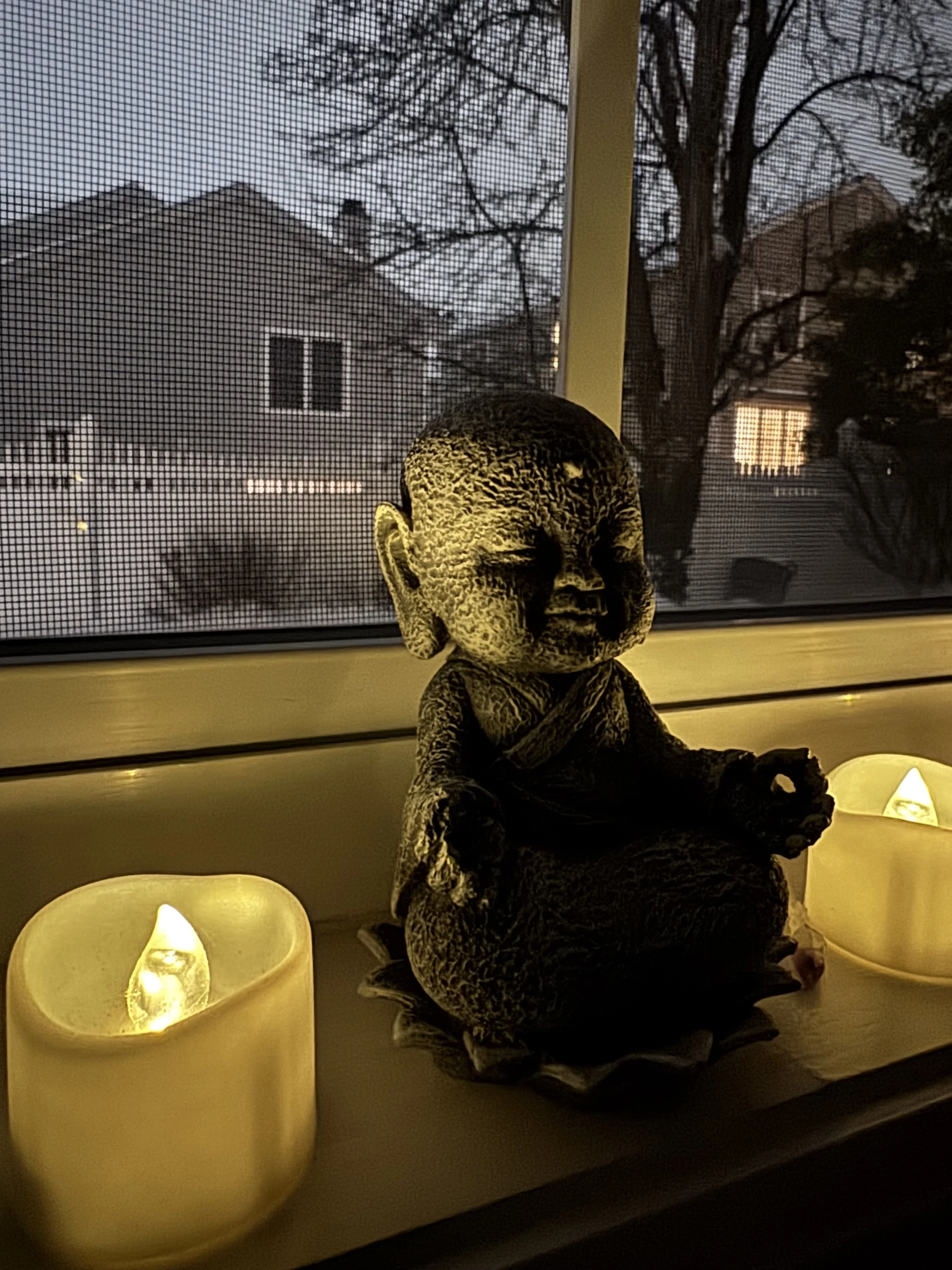 A small figurine of a meditating child monk sits on a windowsill beside two glowing candles, with a mesh screen and houses visible outside in the background.