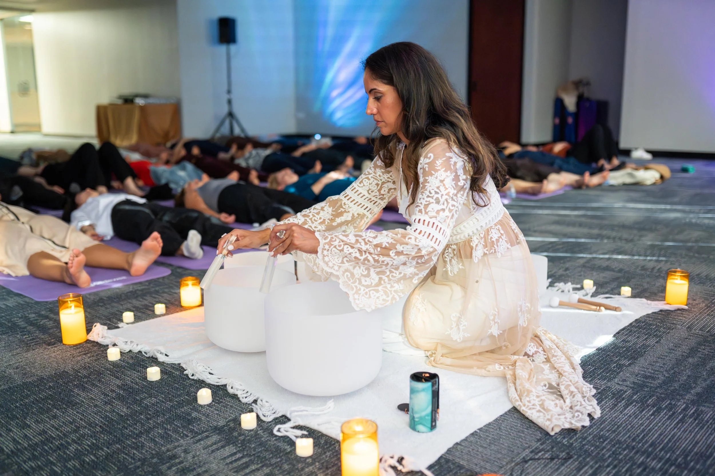 A woman kneeling on a white cloth on the floor, conducting a sound healing session with crystal bowls. People lying down on purple mats in the background during a meditation or wellness class.