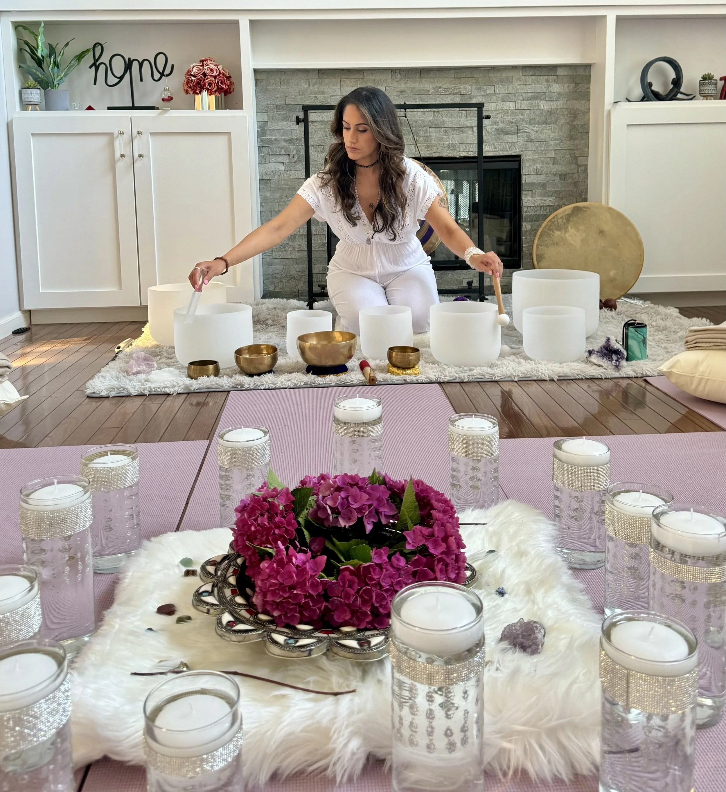 A woman is sitting on a rug in a living room, playing crystal singing bowls during a sound bath session. There are candles and a floral centerpiece on a table in the foreground, with pink hydrangeas and white candles, and the room is decorated with a stone fireplace, shelves, and decorative objects.