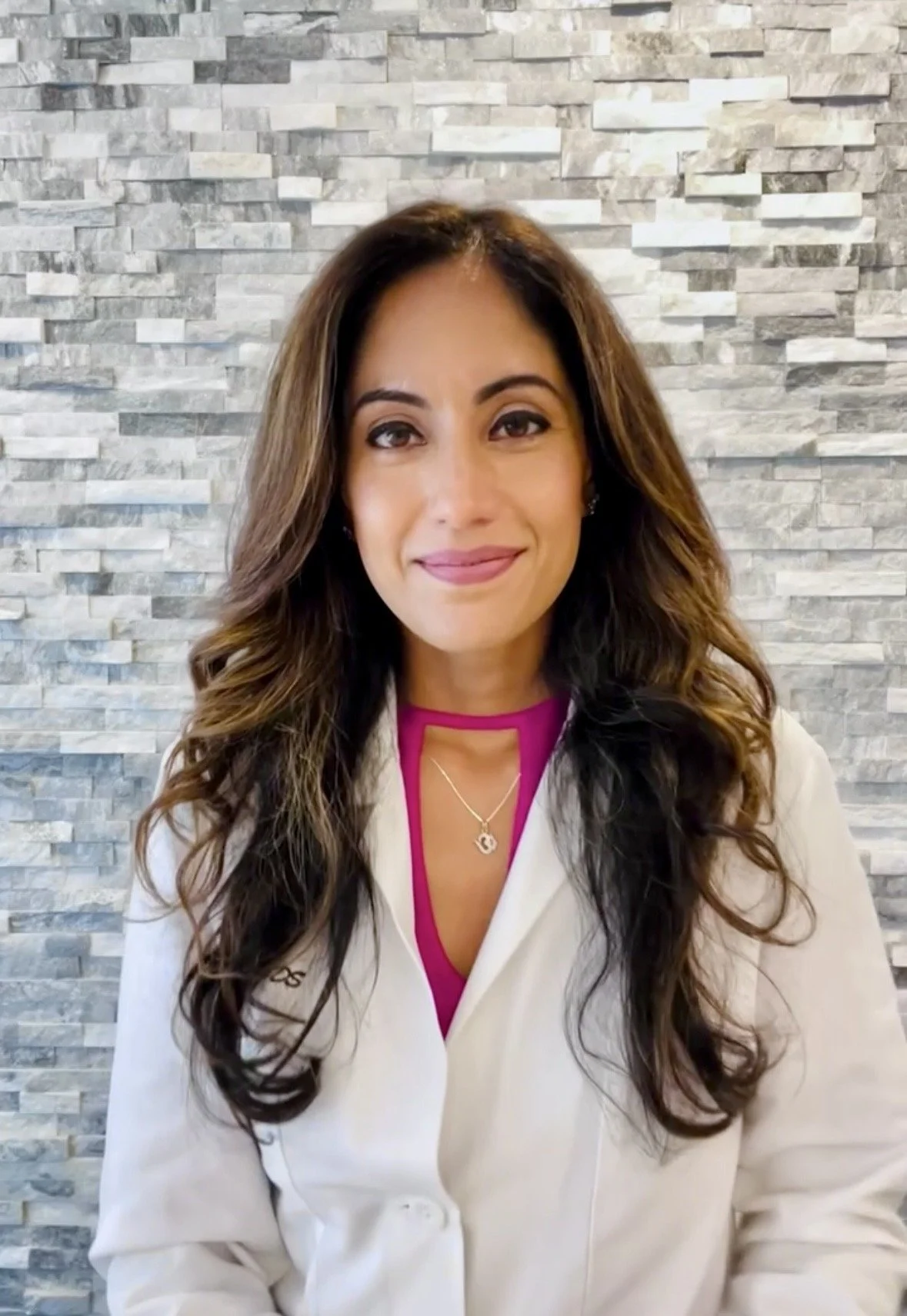 A woman with long, wavy brown hair and a light skin tone smiling at the camera, wearing a white blazer over a magenta top, standing against a gray stone wall background.
