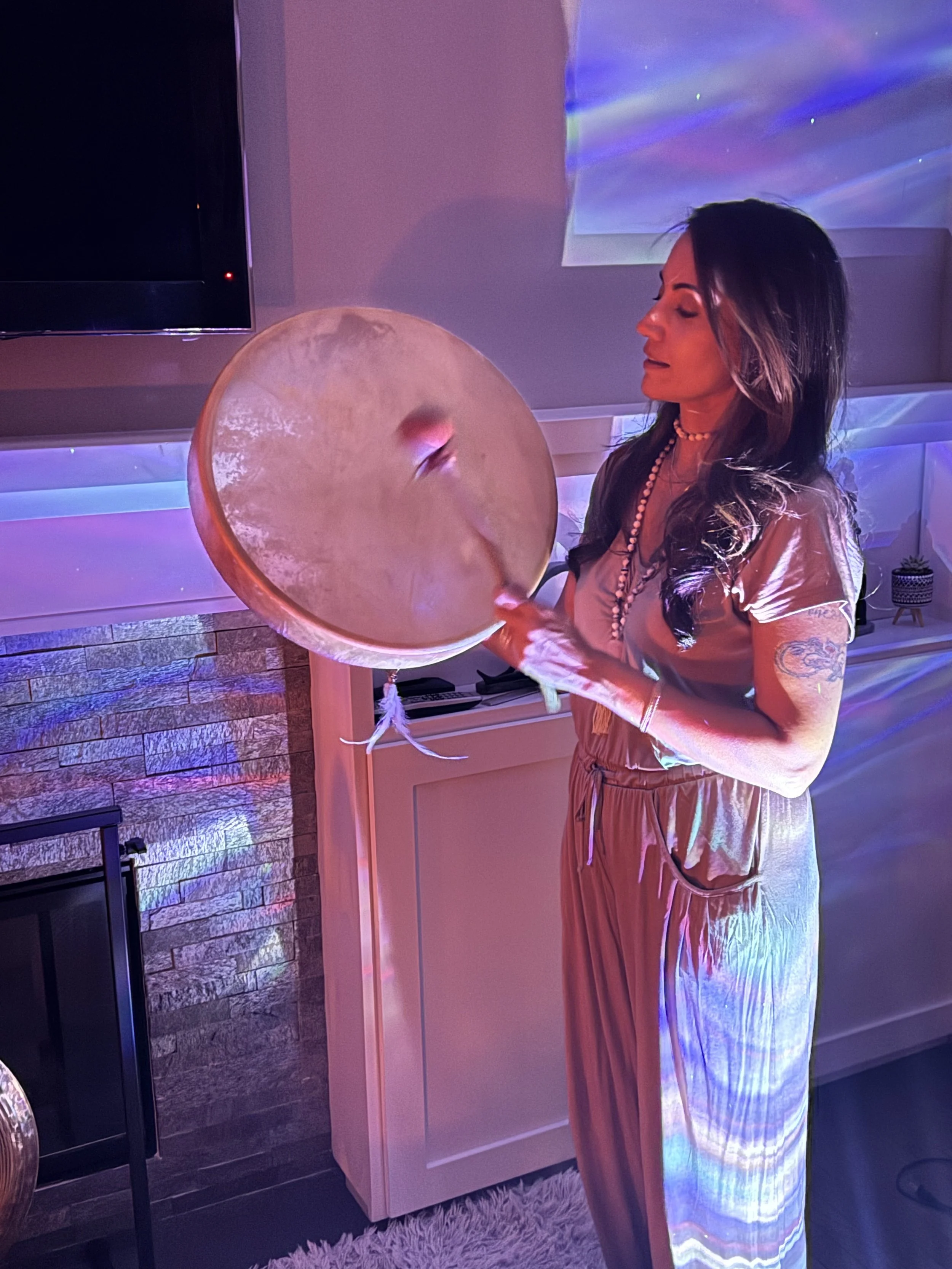A woman with dark hair and tattoos on her arm plays a drum in a room with colorful lights and a TV screen.