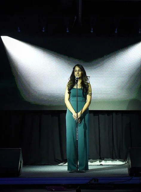 A woman stands on stage in front of a microphone, illuminated by stage lights with a large screen behind her.