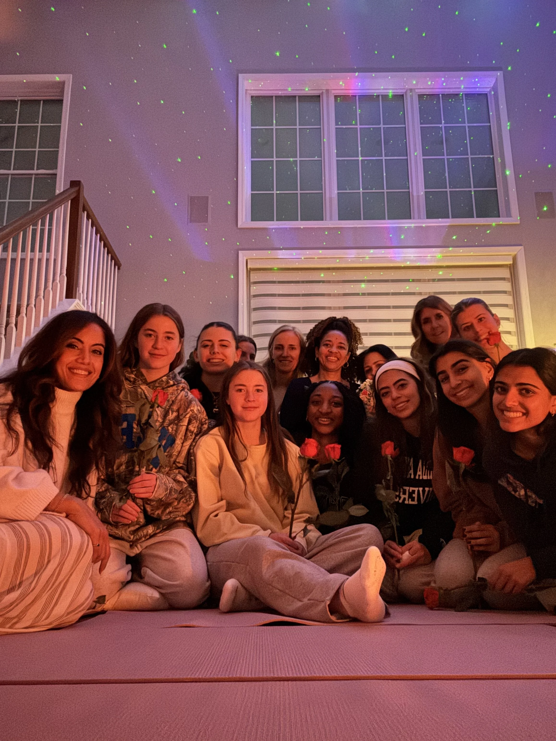 A group of women and girls gathered indoors for a celebration, some holding red roses, with laser lights creating colorful patterns on the ceiling.