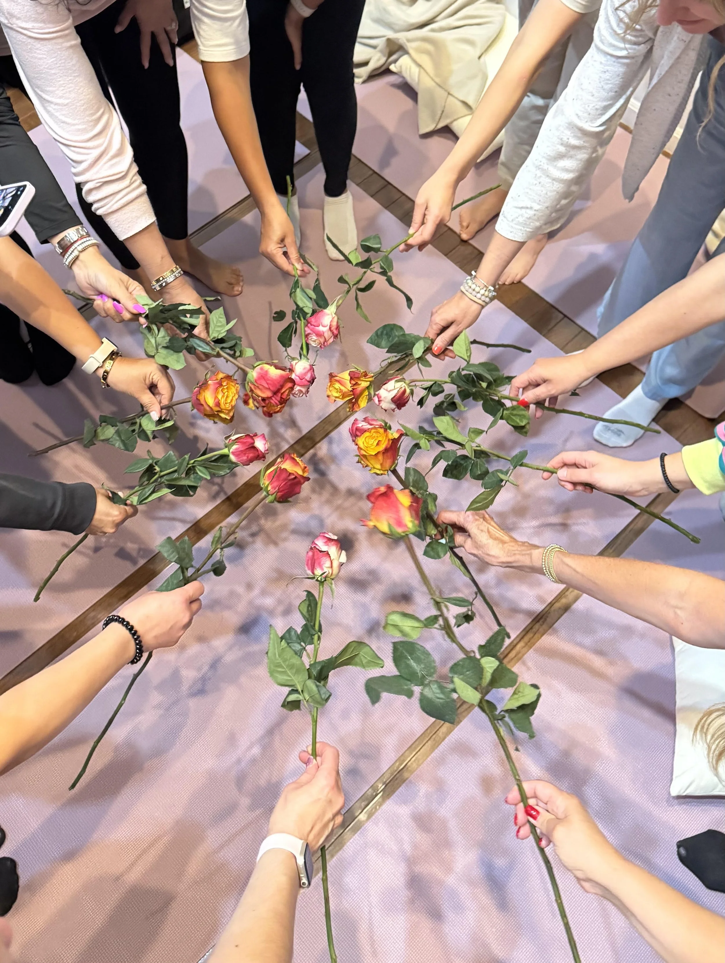 Several people gather around a table, each holding a long-stemmed rose with pink, yellow, and orange blooms, arranged in a circle on the table.