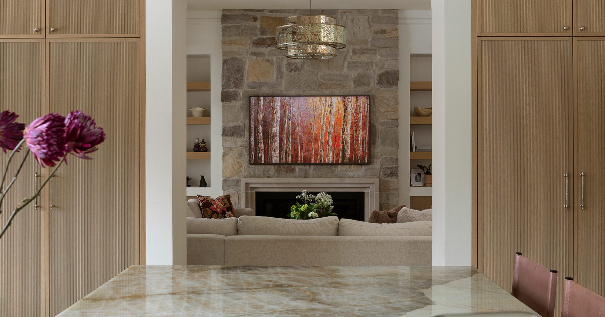 Living room with stone fireplace, wall-mounted TV displaying a forest scene with fall colors, beige sofa with floral and plain pillows, open shelves on either side of the fireplace with decorative items, marble dining table in foreground, pink flower