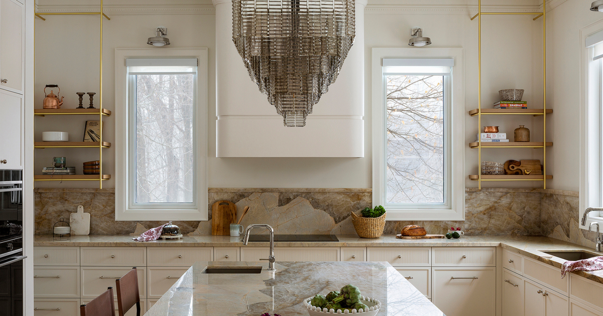 Bright kitchen with two large windows, beige marble countertops, white cabinets, open shelves with decorative items, and a geometric chandelier hanging above the island.