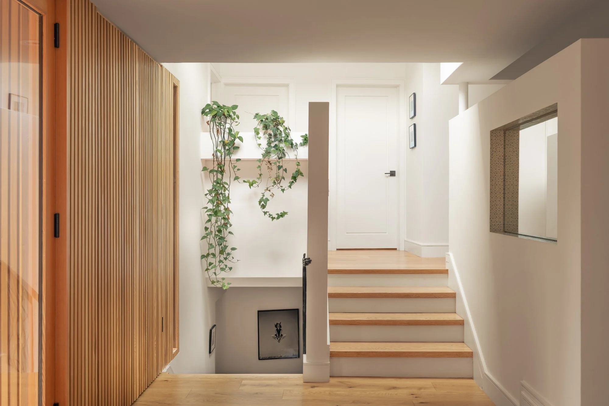 Interior of a modern, minimalist staircase landing with wooden stairs, white walls, a large plant trailing down the wall, and small framed pictures on the wall.