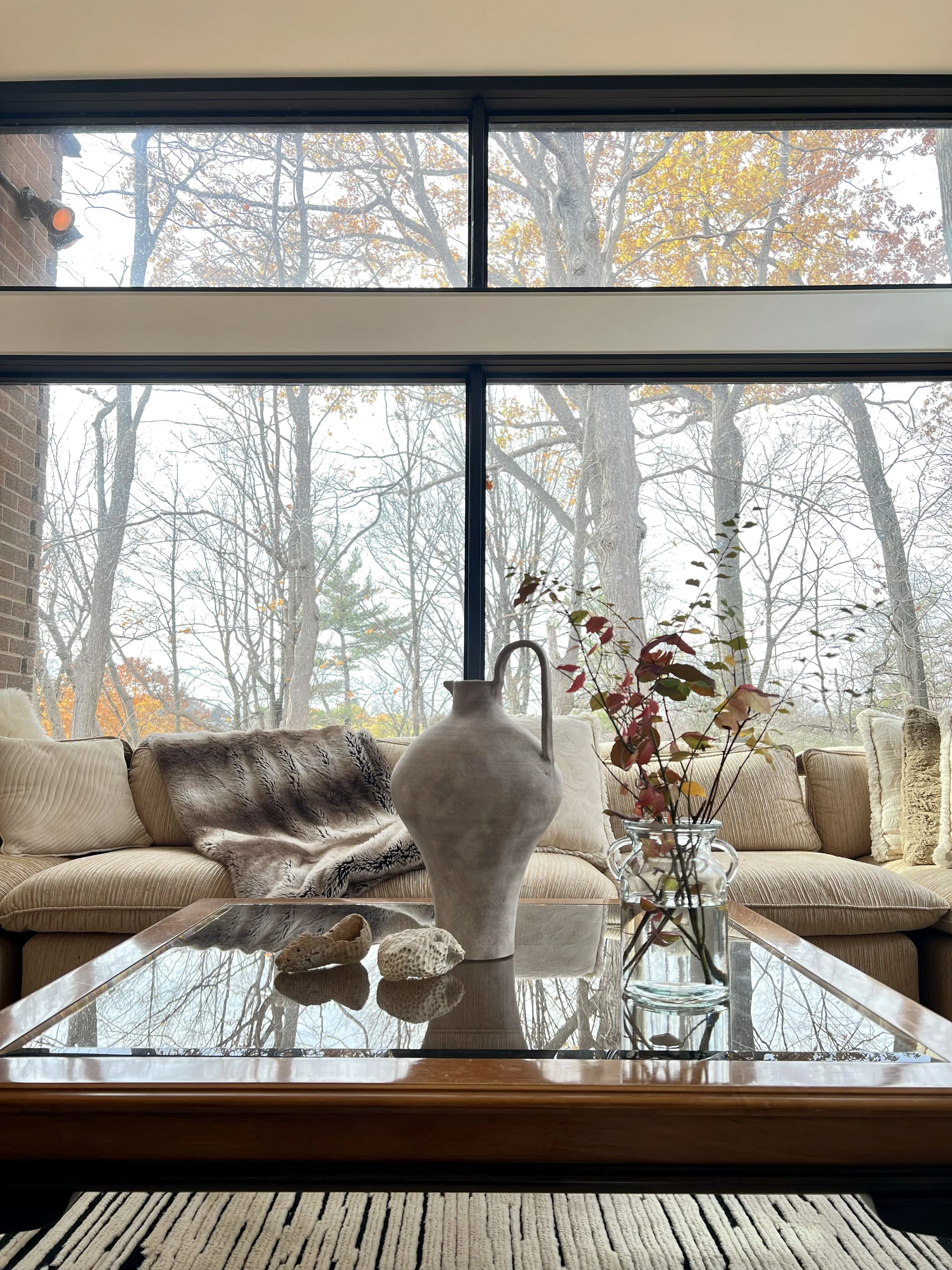 Living room with large windows showing trees outside, beige sofa with pillows, a throw blanket, a coffee table with a vase, decorative rocks, and a glass jar with branches and leaves.