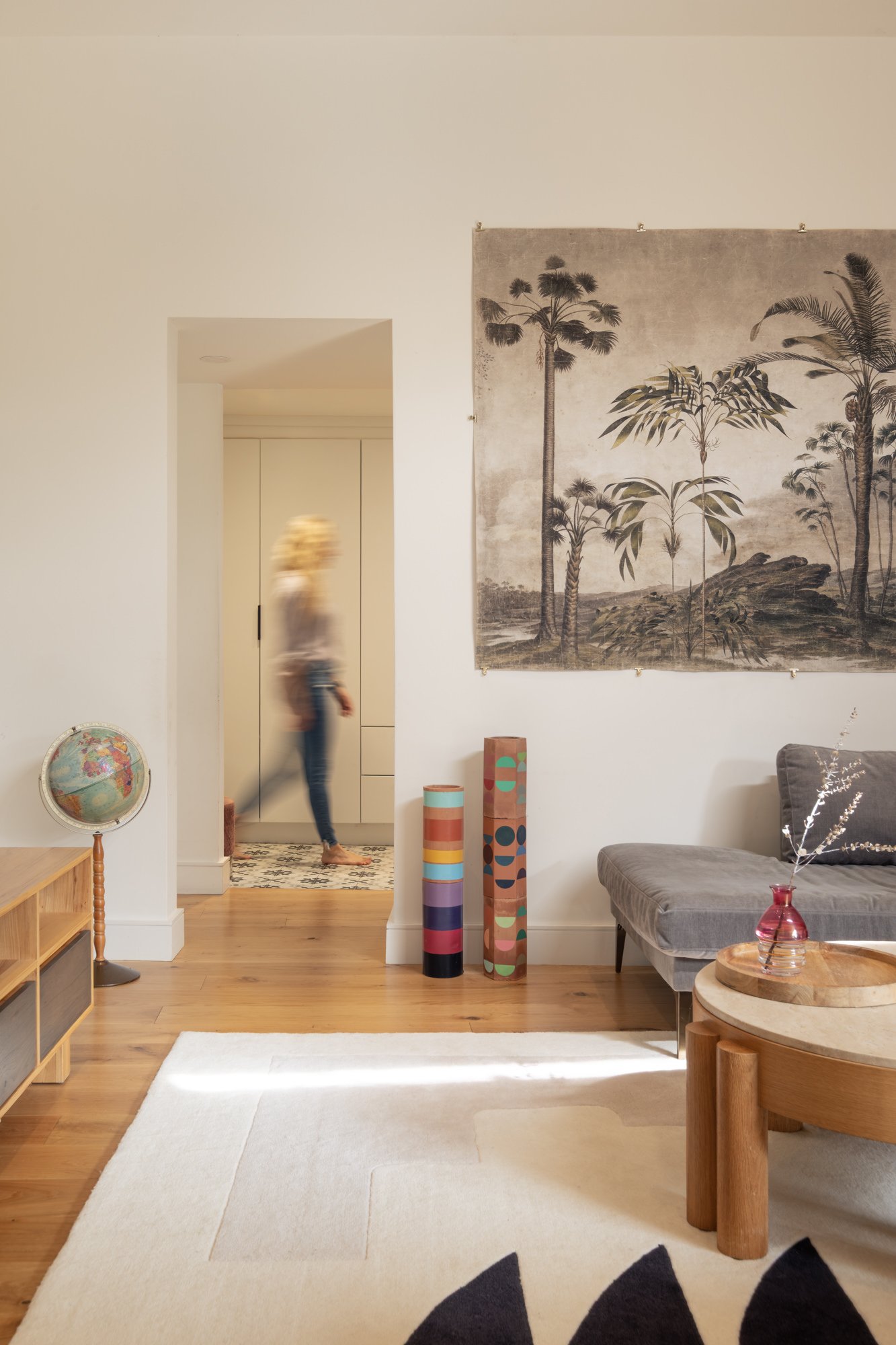 A cozy living room with a wooden floor, a white rug, and a gray sofa. On the wall, there's a large black-and-white and sepia-toned tropical landscape tapestry. Near the entrance to another room, there are two colorful cylindrical decorative objects a