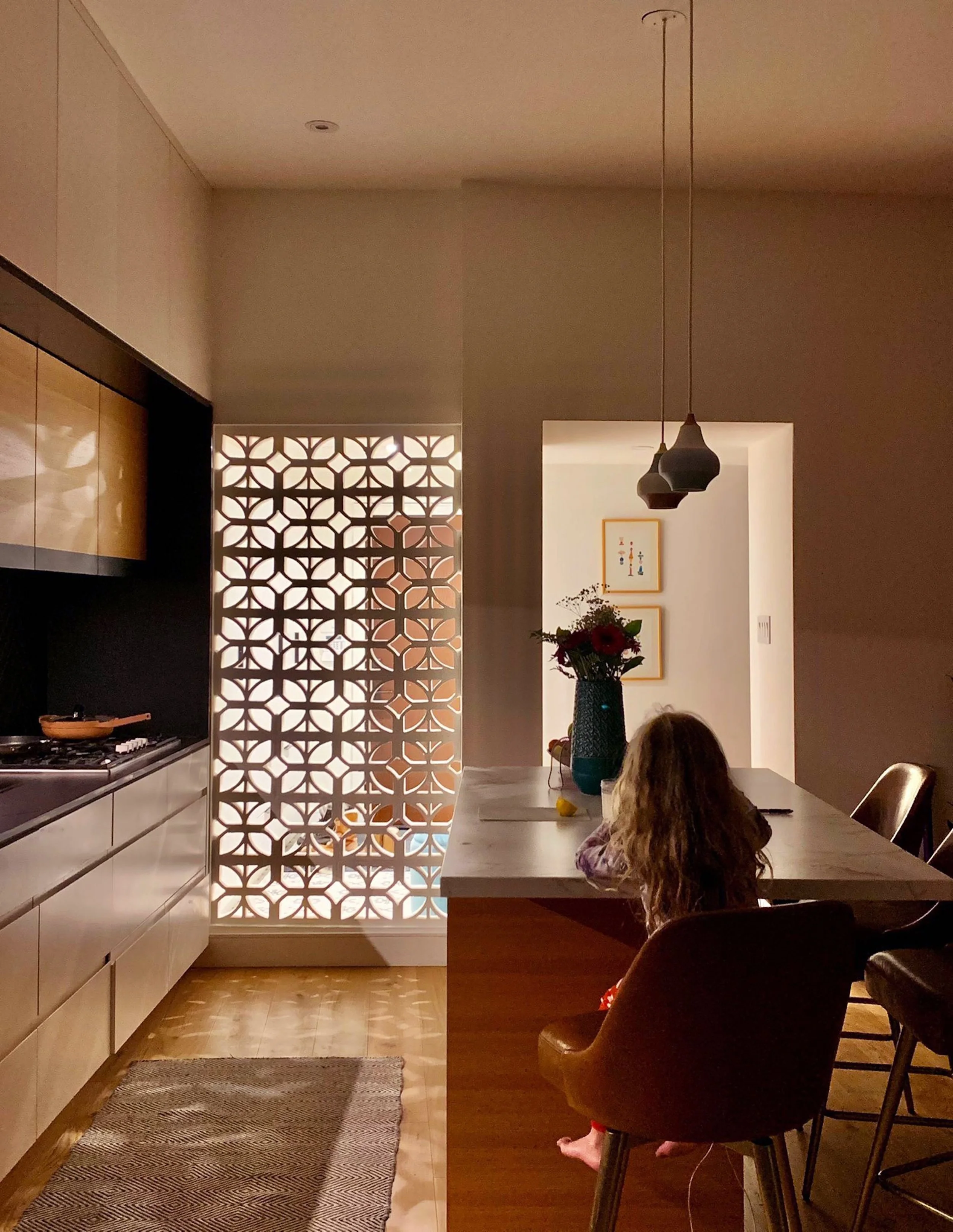 A kitchen with a white counter, a decorative wall divider with geometric patterns, and a dining area with a large table, a vase of flowers, and a young girl with long curly hair sitting in a chair.