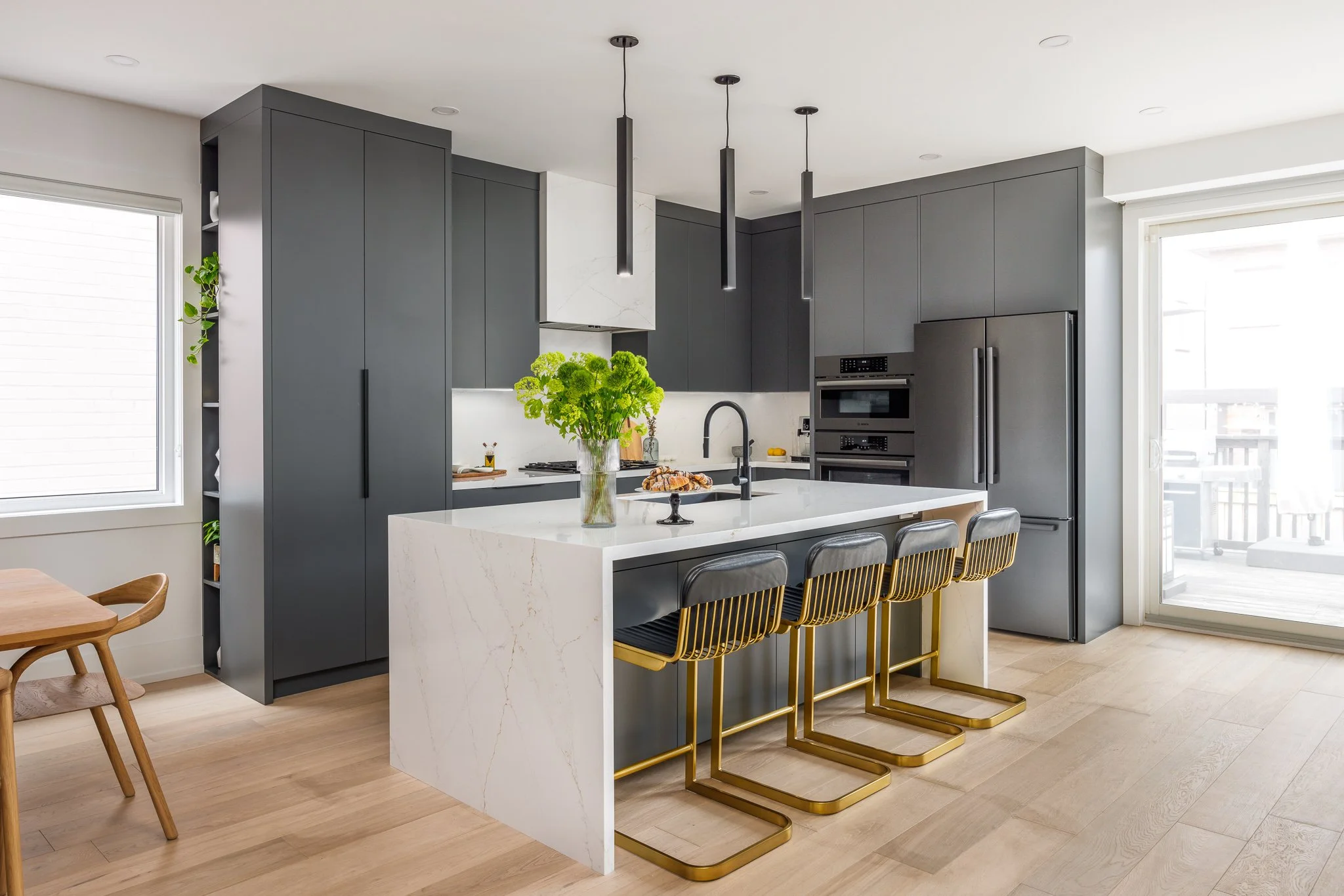 Modern kitchen with dark gray cabinets, white marble island with black stools, and a sliding glass door leading to a balcony.