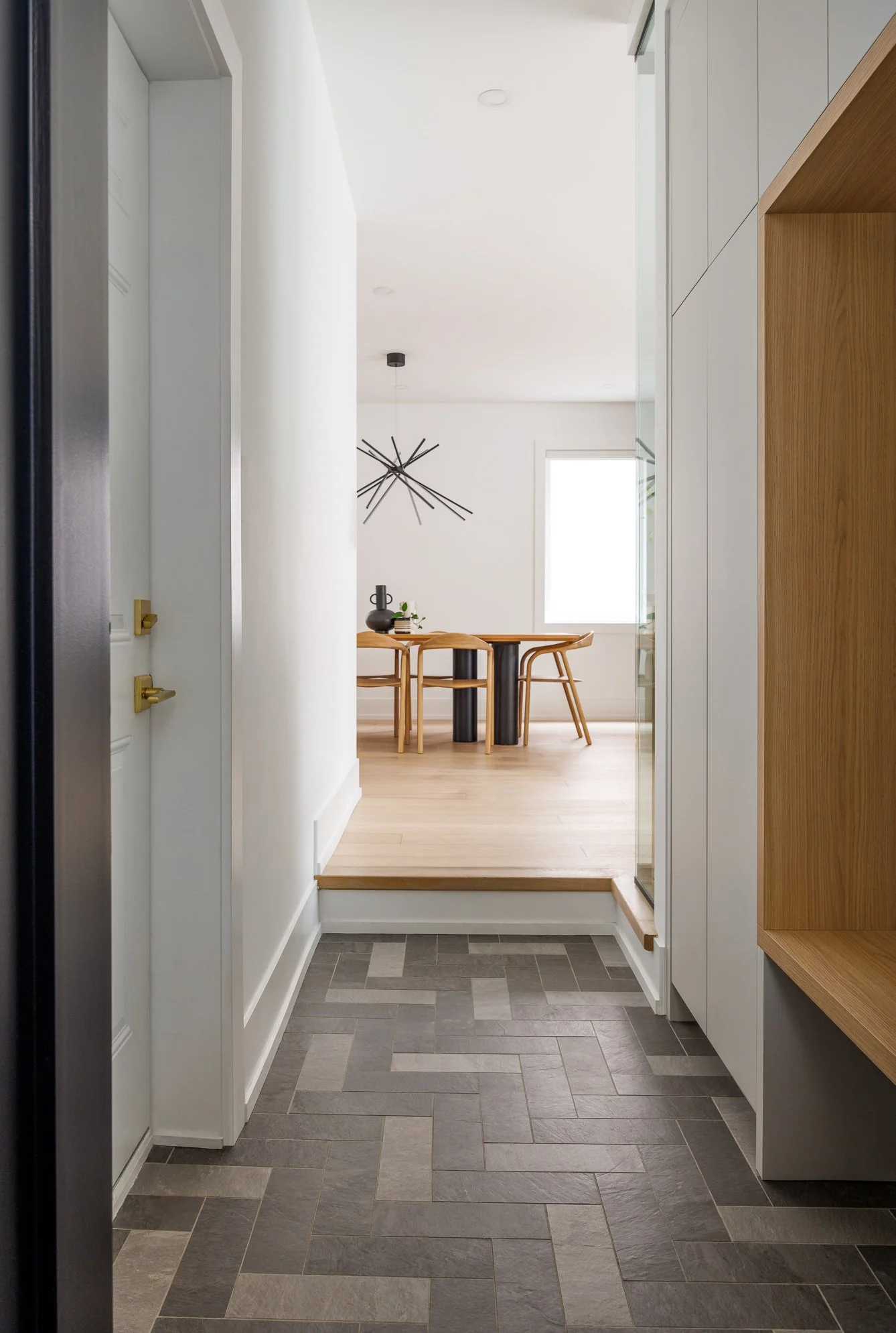 Entryway view into a dining area with a modern chandelier, a wooden dining table, and chairs, with a large window in the background.