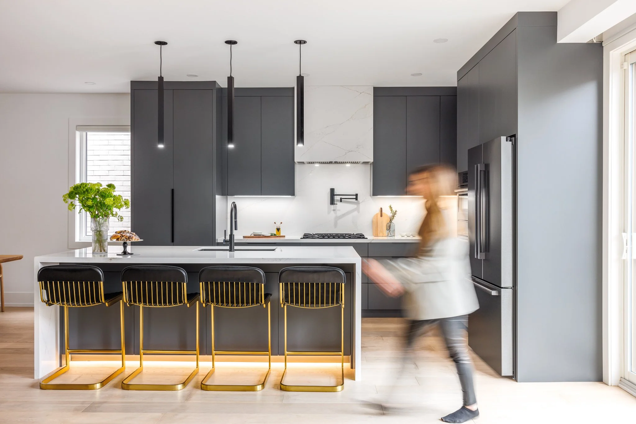 Modern kitchen with black cabinets, white marble island, black and gold bar stools, and a woman walking past.
