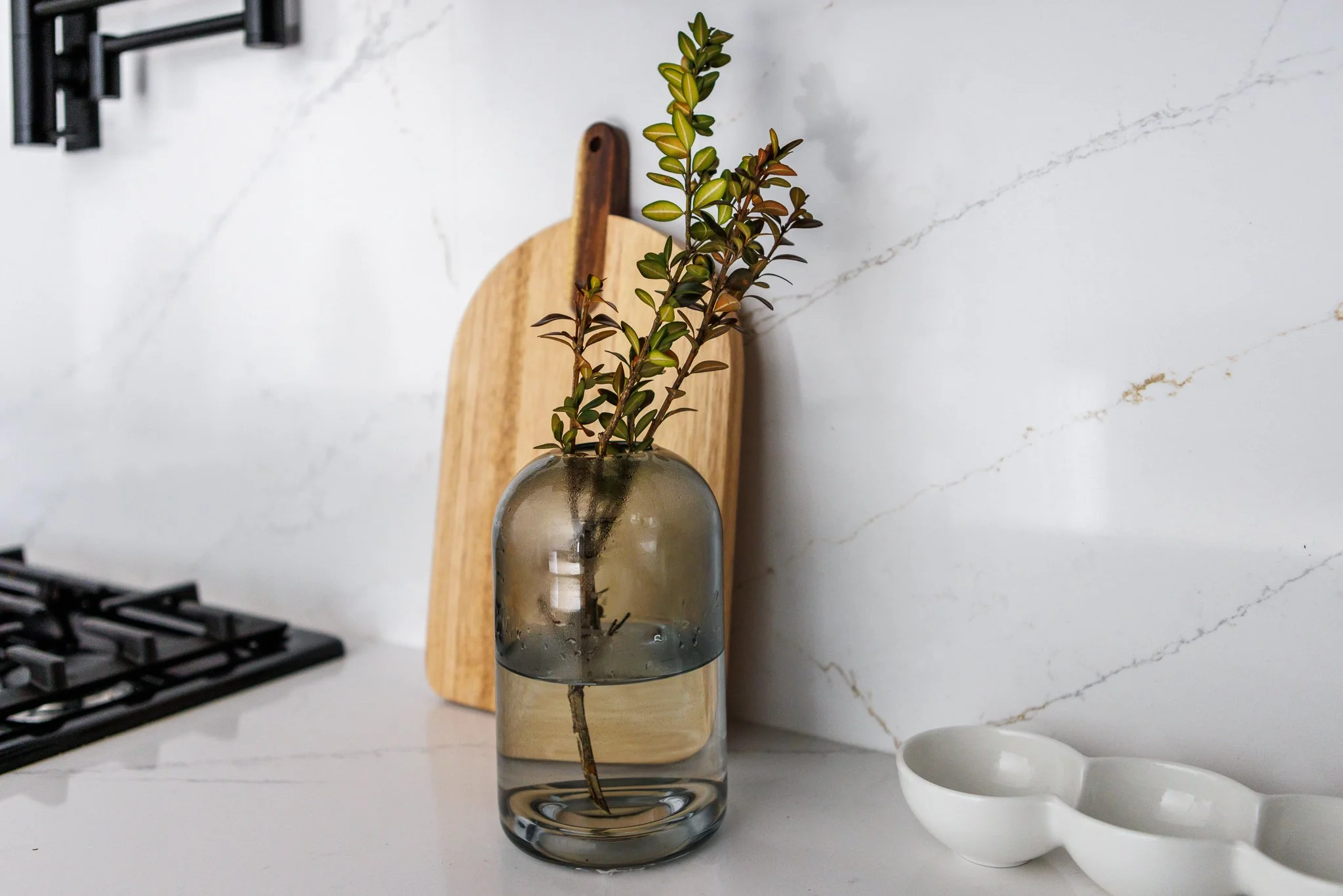 A kitchen countertop with a glass vase holding a plant, a round wooden cutting board with a handle, and a white ceramic dish with two compartments. Part of a black stovetop is visible on the left side. The background features a white marble wall with