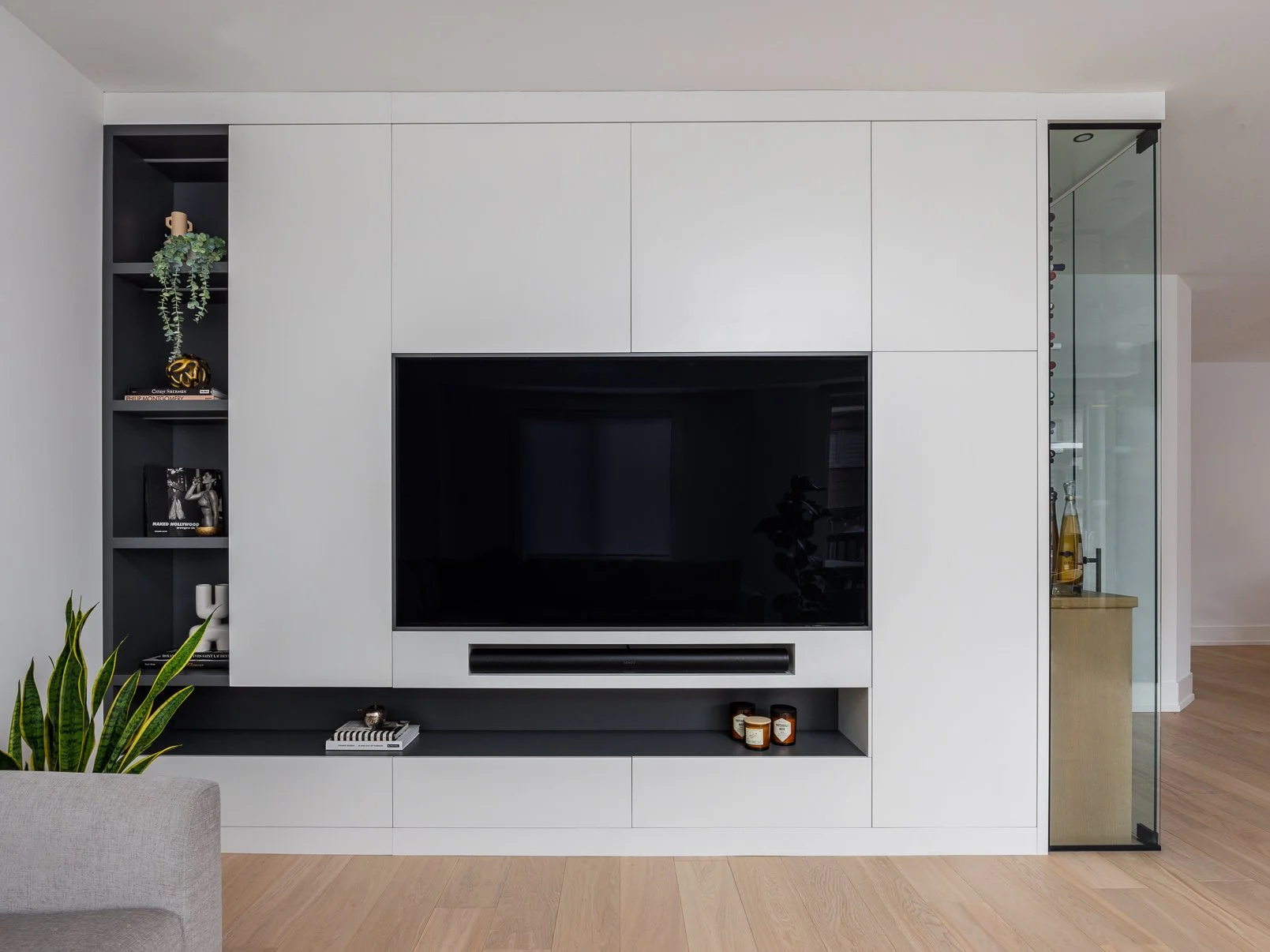 Modern living room entertainment center with a large flat-screen television, open shelving with books and decor, a plants on the left, and a glass door on the right revealing bottles inside a cabinet.